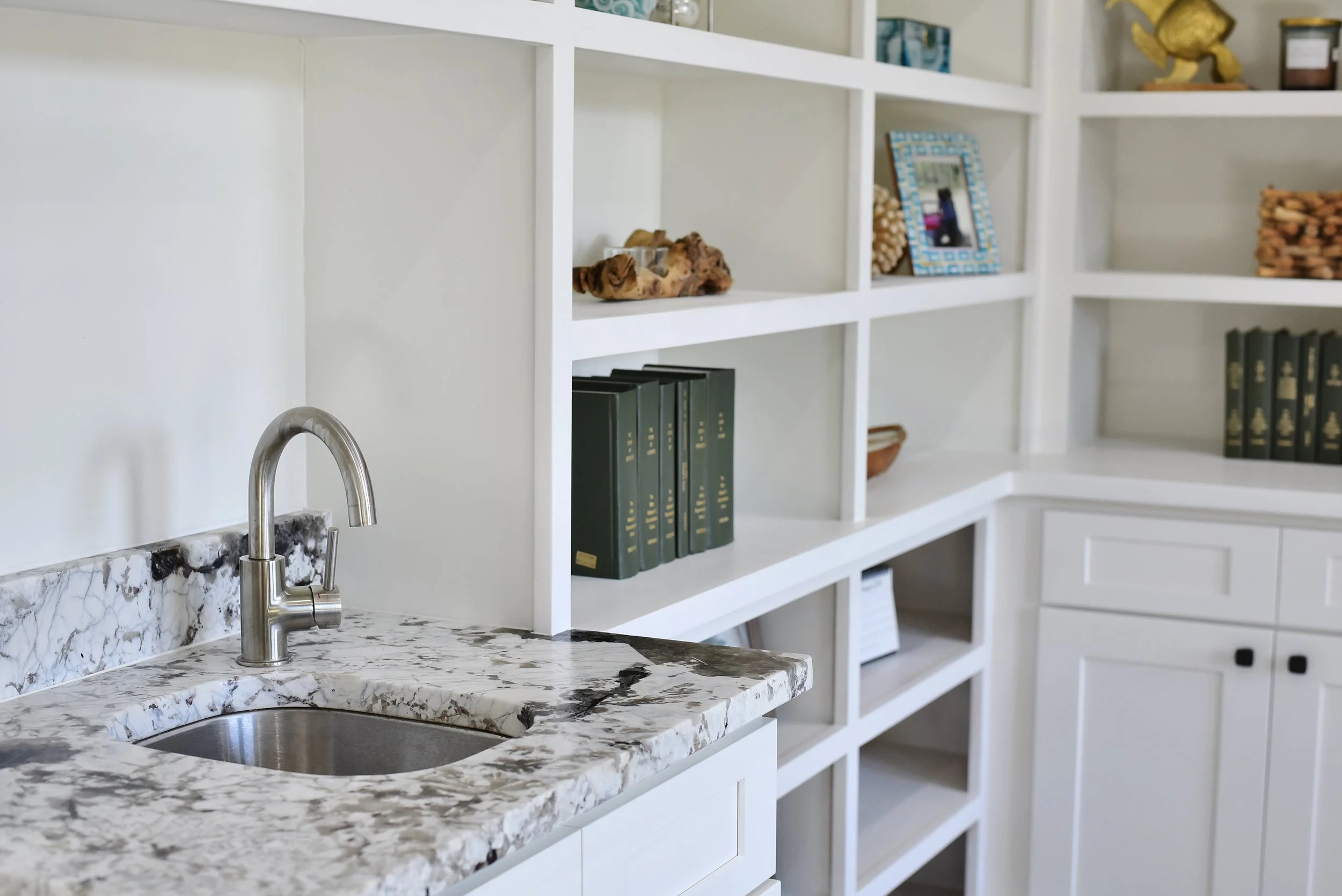 White kitchen with marble countertop sink, stainless steel faucet, and white shelving filled with books, decorative items, and framed photos.