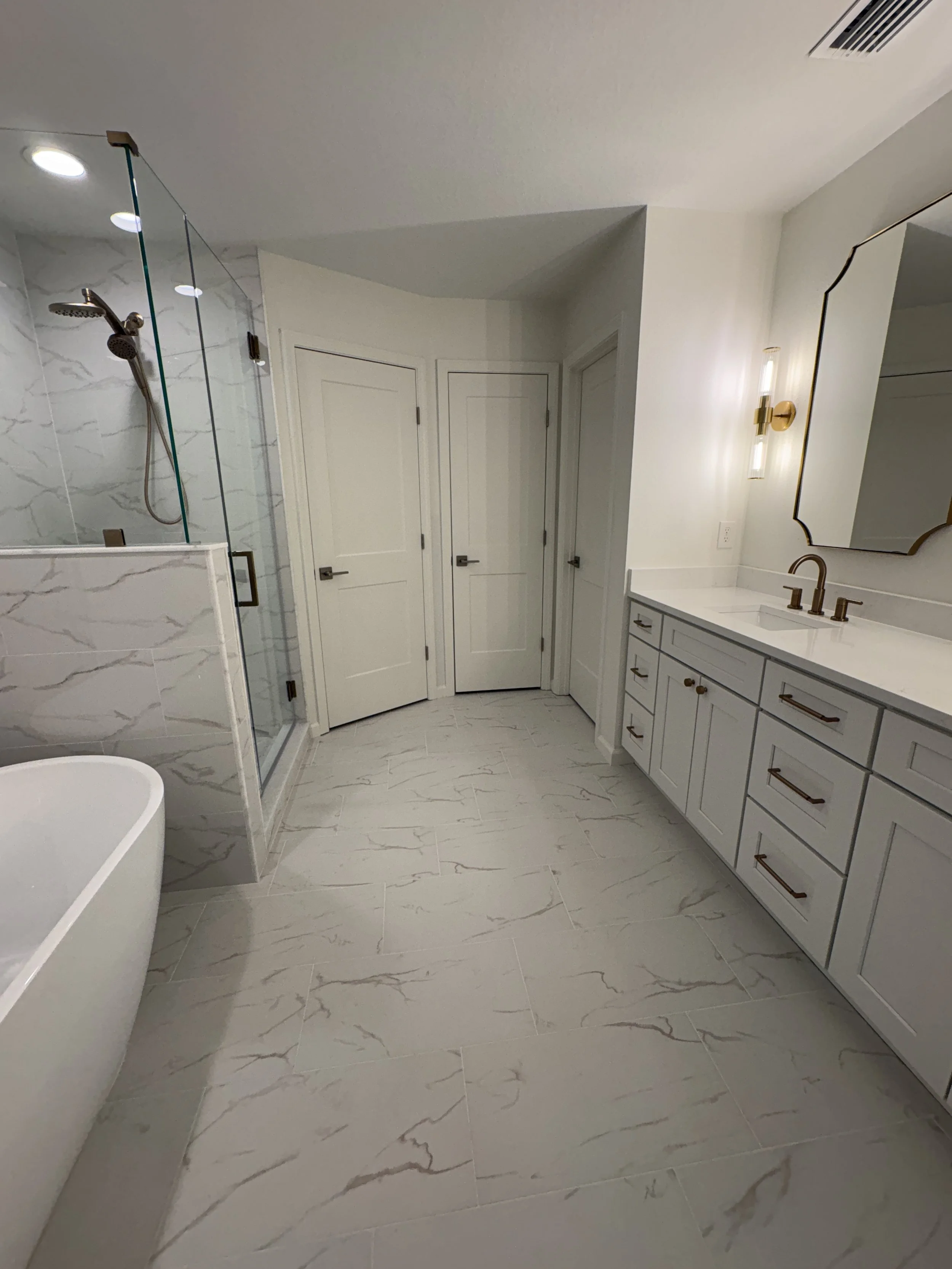 This completed luxury primary bathroom combines a custom double vanity, frameless glass shower, freestanding tub, marble-look tile, and brushed gold fixtures. Elite Construction of JAX completed this high-end full-condo remodel in Jacksonville, Flori