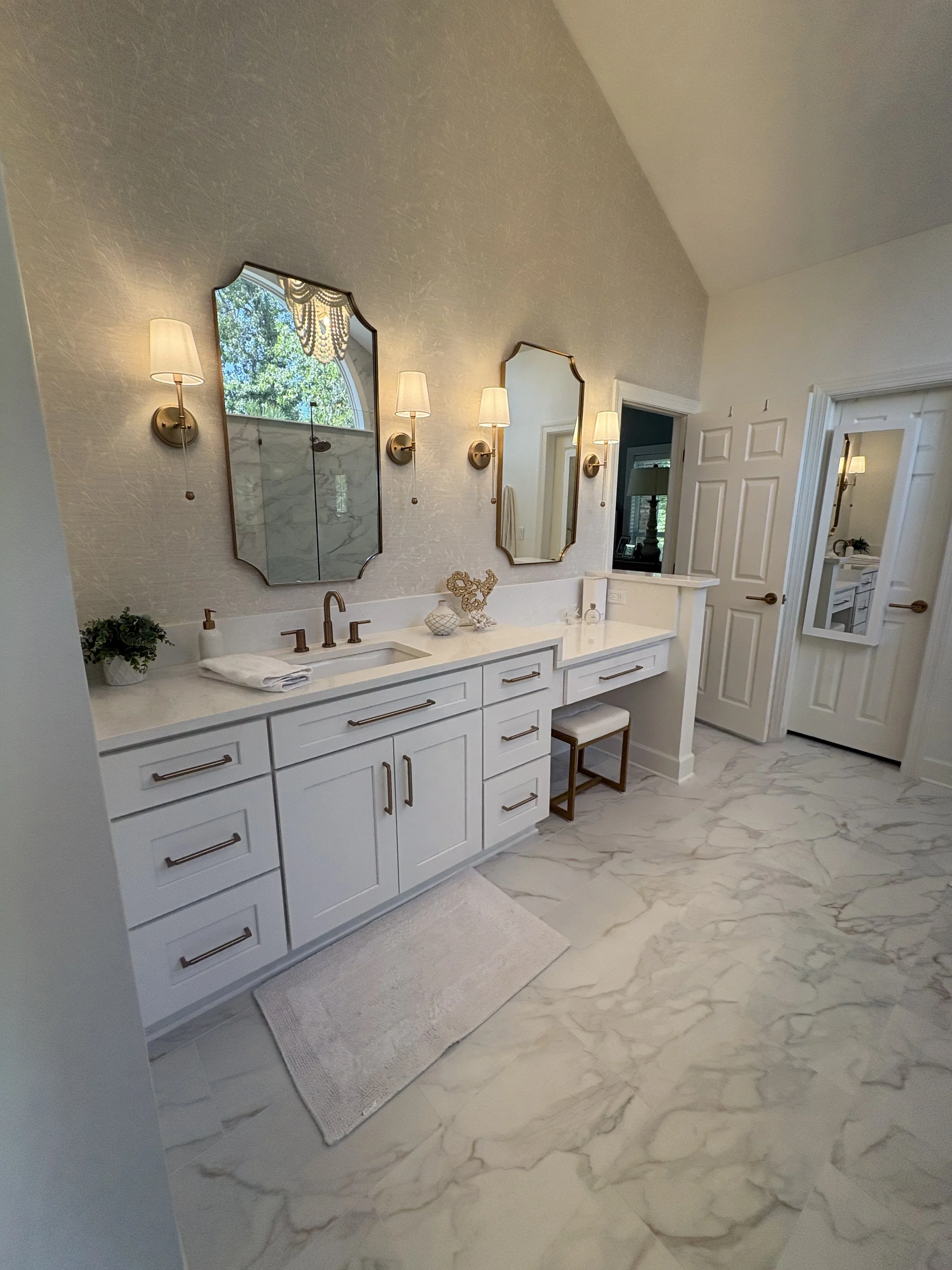 Finished primary bathroom featuring a custom double vanity with white shaker cabinetry, quartz countertops, and a built-in makeup station designed for both style and functionality. Decorative mirrors, elegant wall sconces, and expansive marble-look p