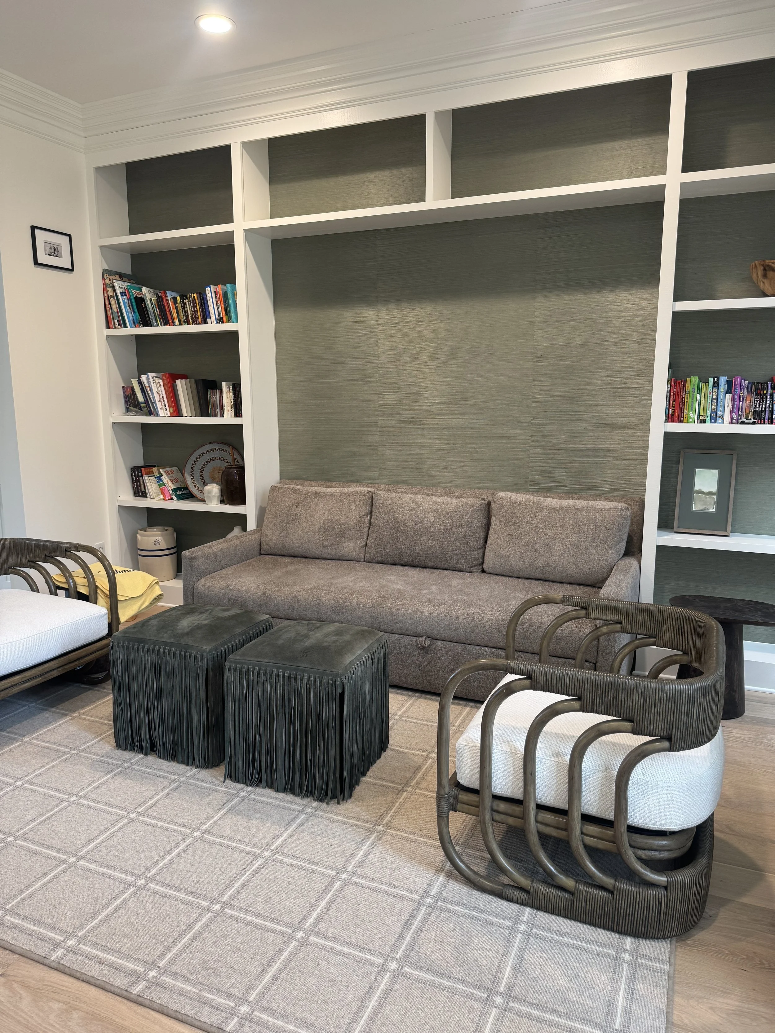 Living room with a gray couch, two dark green ottomans, and two armchairs with brown curved frames and white cushions. Behind the couch is a built-in white bookshelf filled with books and decorative items. There is a beige rug with a checkered patter