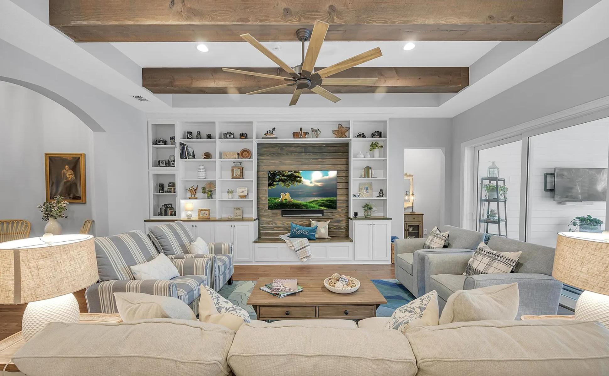 Living room with beige and gray sofas, wooden ceiling beams, a ceiling fan, and a built-in entertainment center with a TV and white shelving, decorated with books and ornaments.