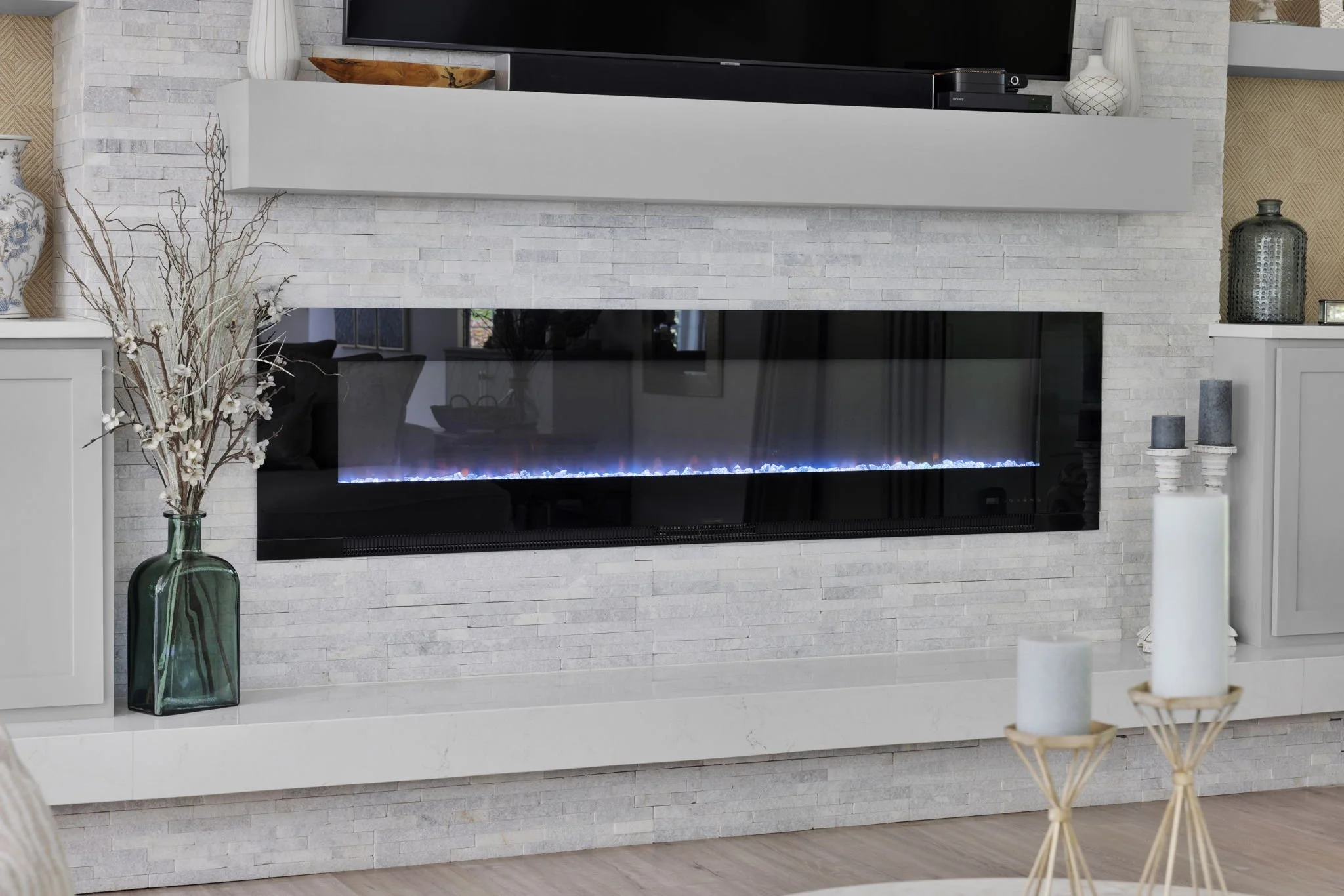 Living room fireplace with white textured brick wall, black glass front, and a TV mounted above. Decor includes vases, candles, and a glass jar with branches.