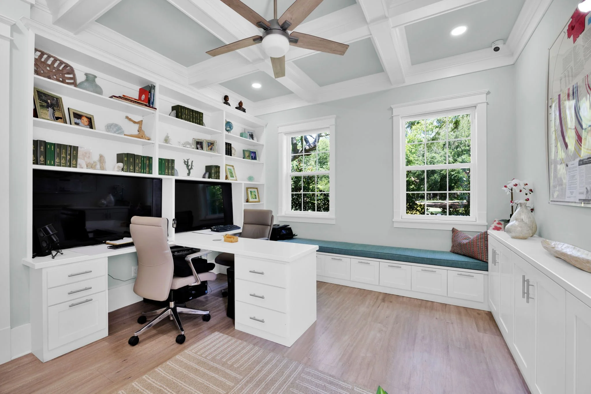 A modern home office with white built-in shelves and cabinets, two computer monitors on a white desk, beige office chair, two windows with greenery outside, a ceiling fan, and decorative items including vases and framed photos.