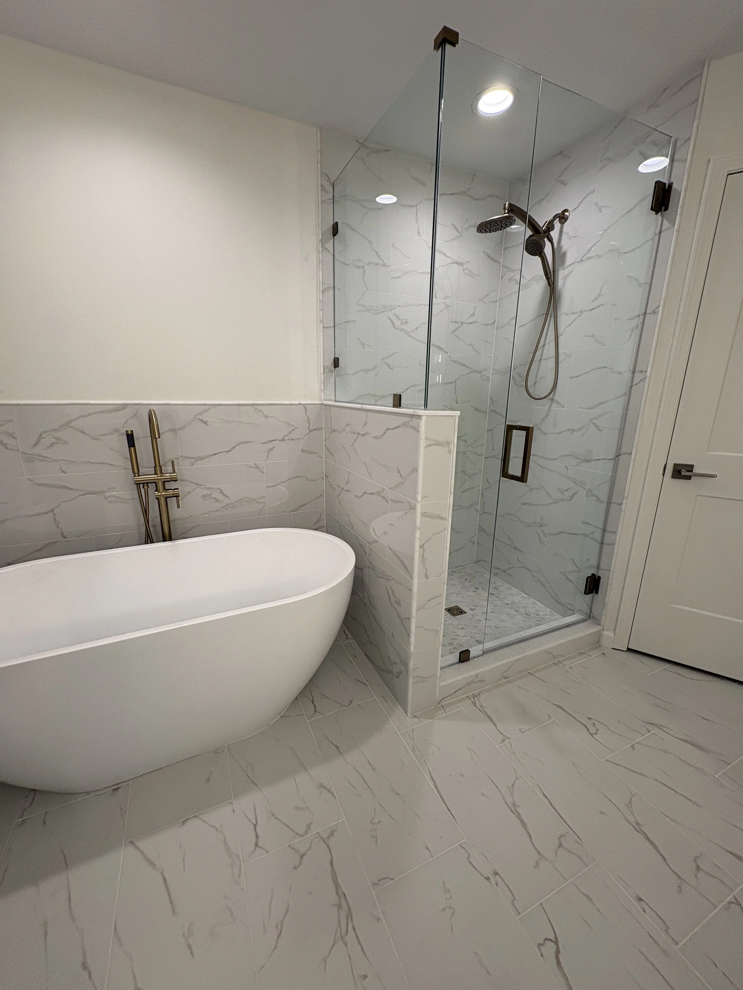 This luxury primary bathroom remodel features a sculptural freestanding tub, frameless glass shower, marble-look tile, and brushed gold fixtures. Elite Construction of JAX completed this high-end condo renovation in Jacksonville, Florida.