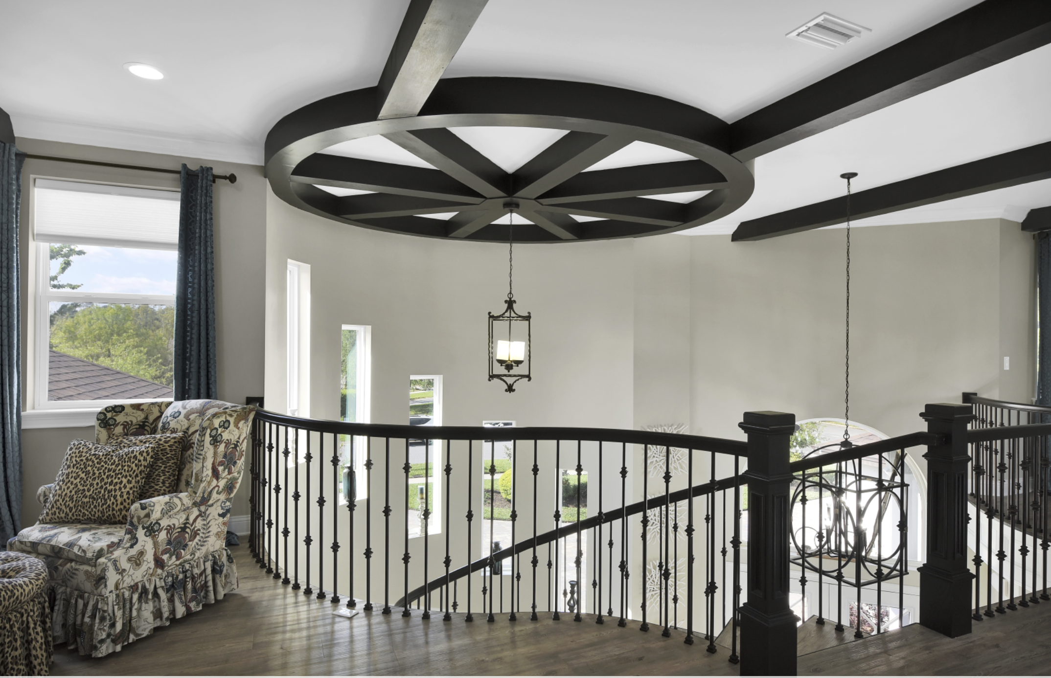 Interior view of a modern home staircase landing with a black wrought iron railing, a floral armchair with leopard print pillows, three small windows, and a decorative ceiling feature with a hanging light fixture.