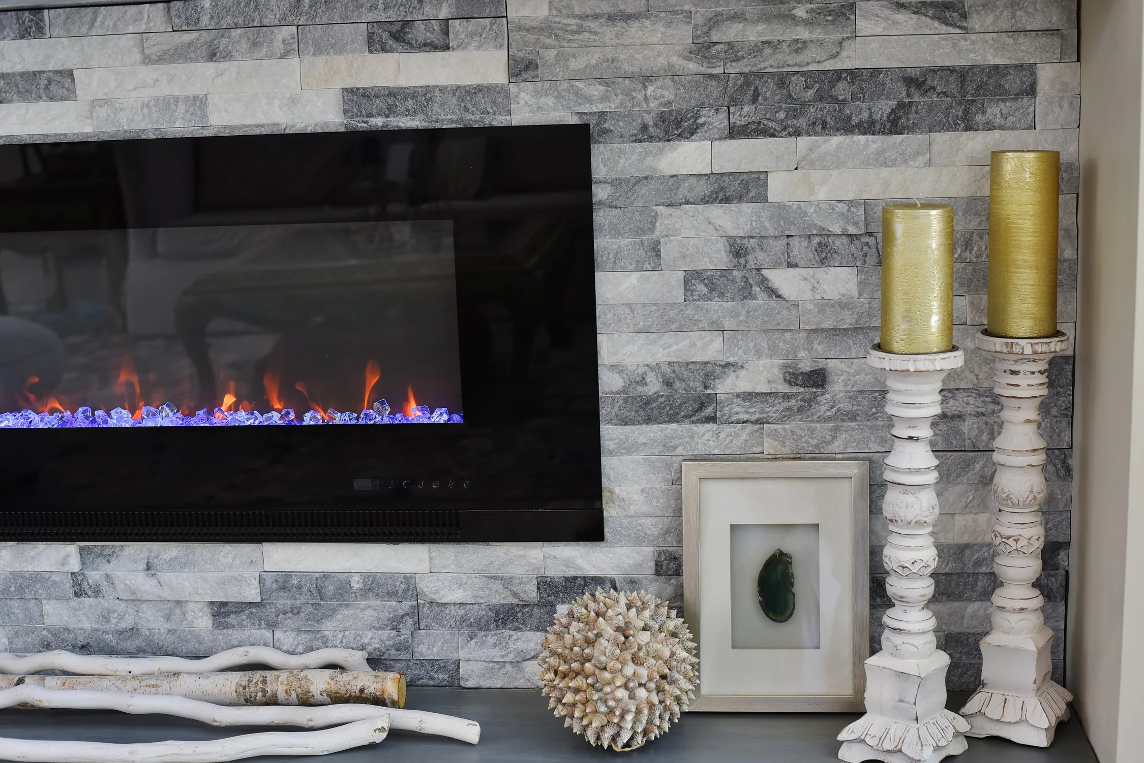 Decorative fireplace with a stone wall, two white candlesticks with gold candles, a framed green stone, a sea urchin shell, and white driftwood branches.