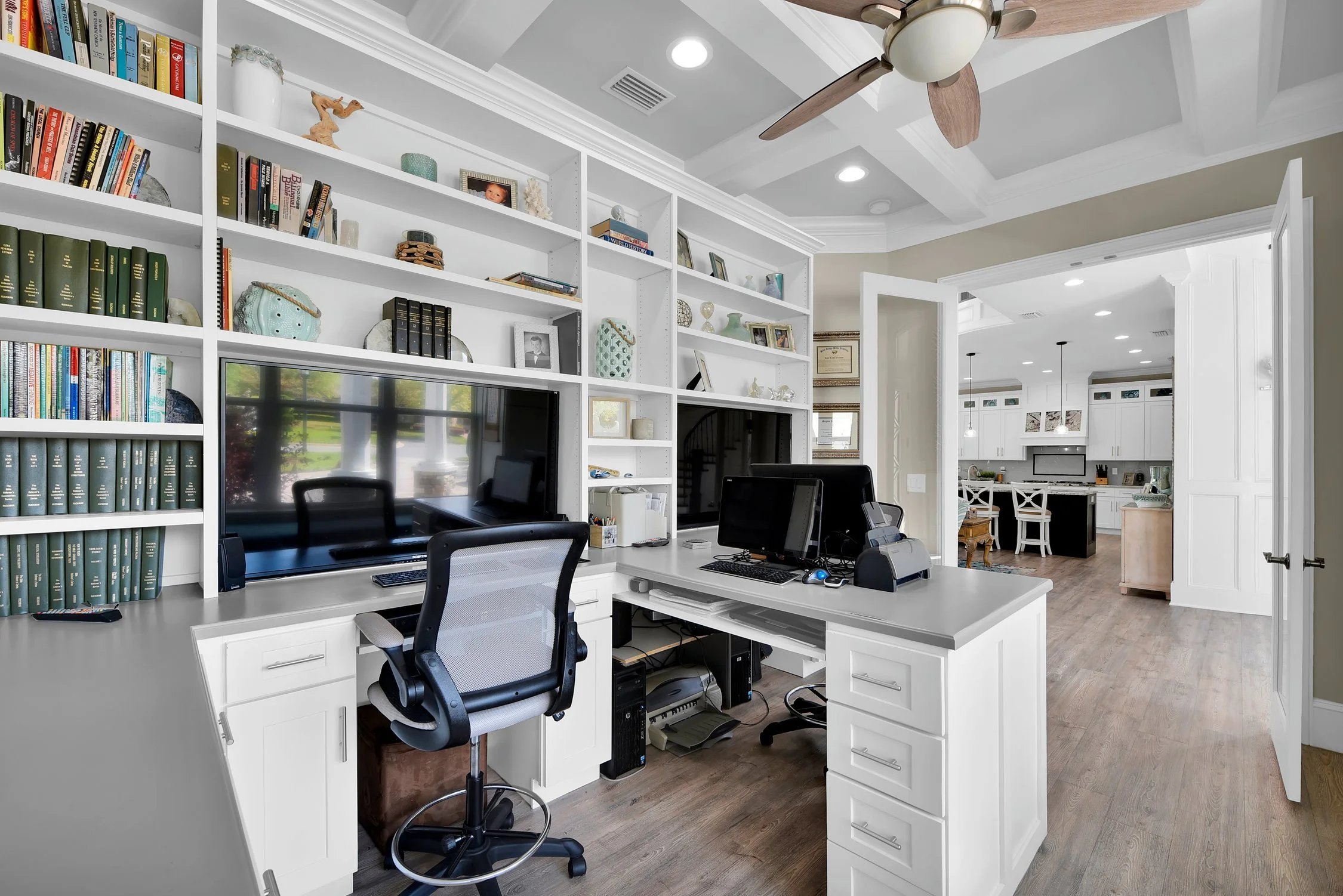 Home office with white desk and shelves, two monitors, office chair, and computers, with a view into a kitchen in the background.
