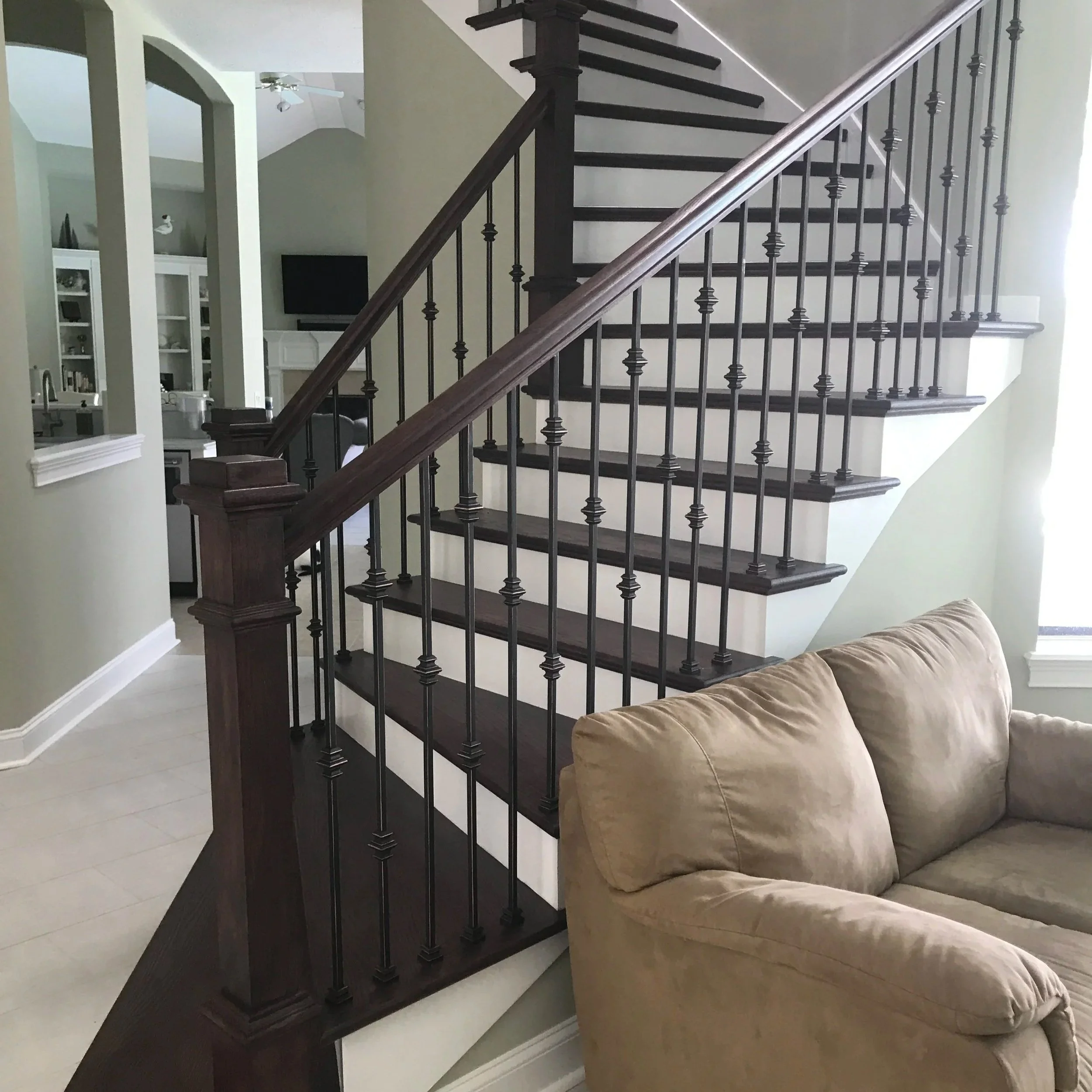 Interior of a home with a staircase featuring dark wood steps and black metal balusters, beige walls, a beige sofa, and a view into the kitchen and living area.