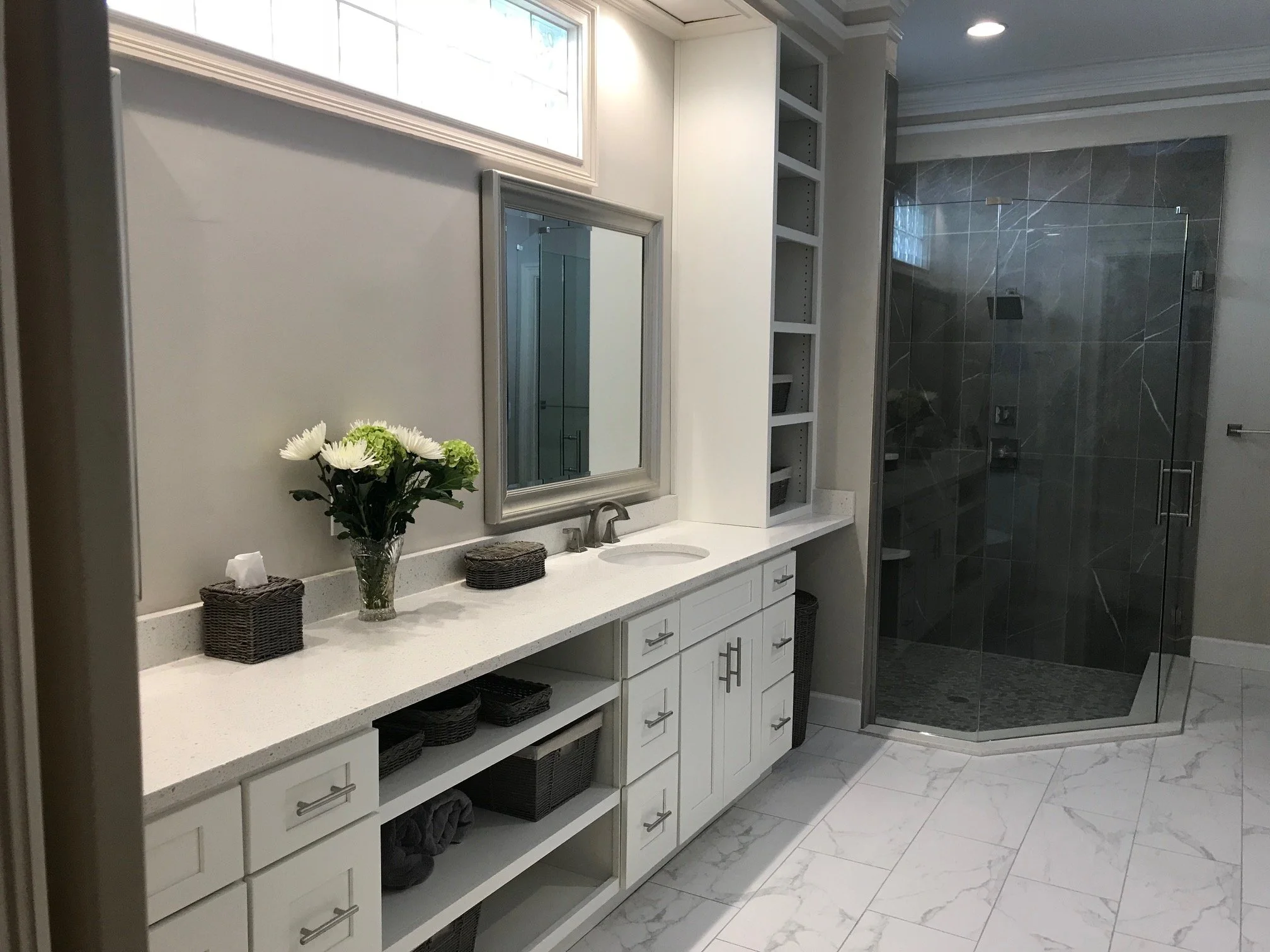 Modern bathroom with white vanity, mirrored cabinets, a glass enclosed shower with dark gray tiles, and decorative elements like a flower vase and woven baskets.
