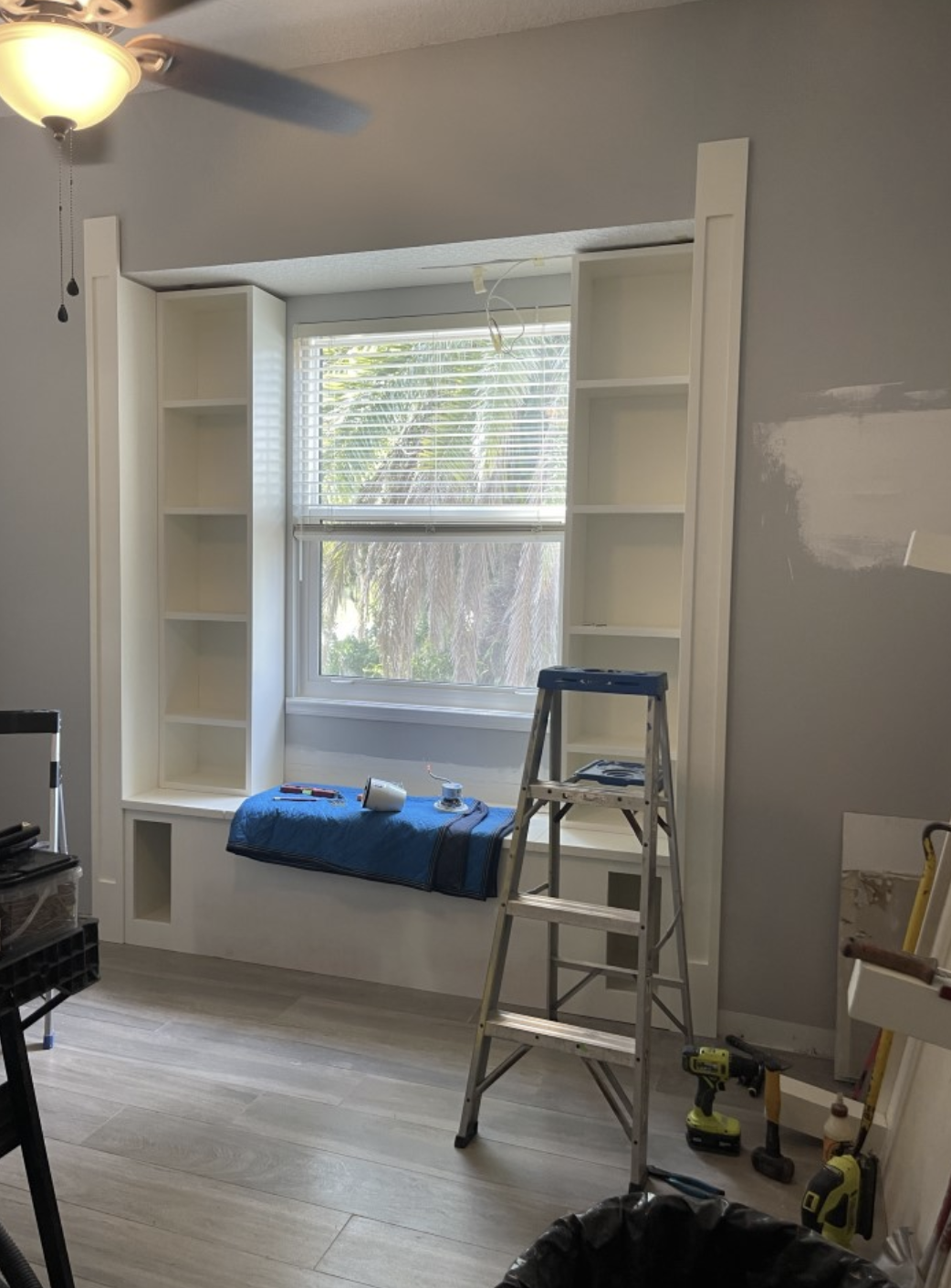 Room under renovation with built-in white bookshelves around window, a step ladder, power drill, and tools on the floor.