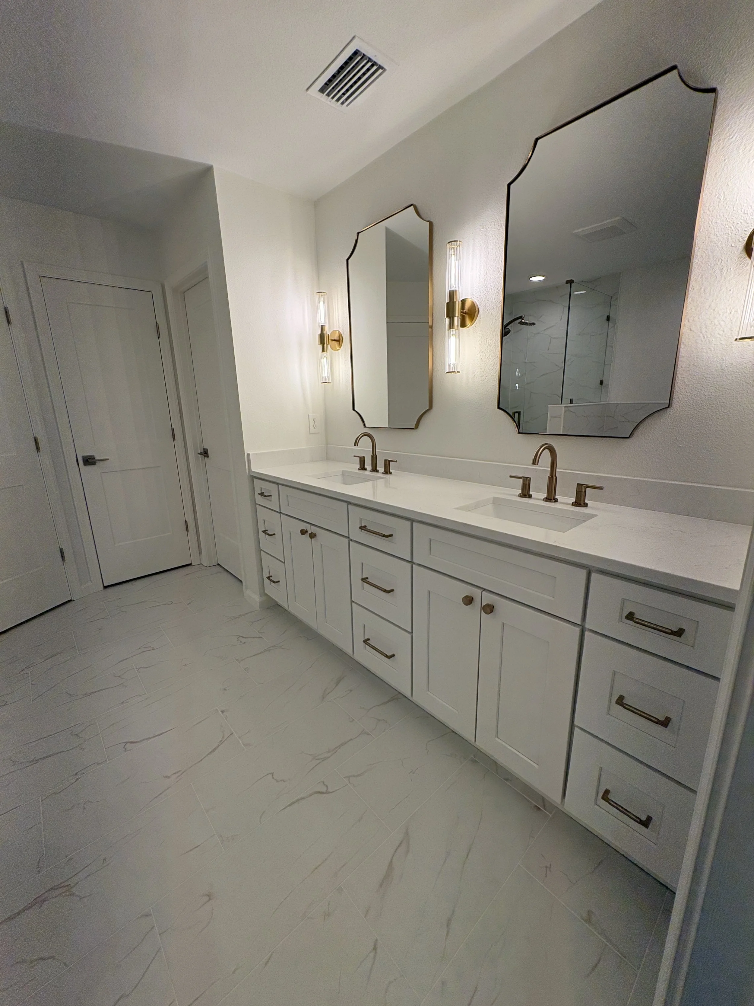 A custom double vanity with extensive storage, a sleek white countertop, decorative mirrors, and brushed gold fixtures brings elegance and function to this luxury Jacksonville condo bathroom remodel by Elite Construction of JAX.