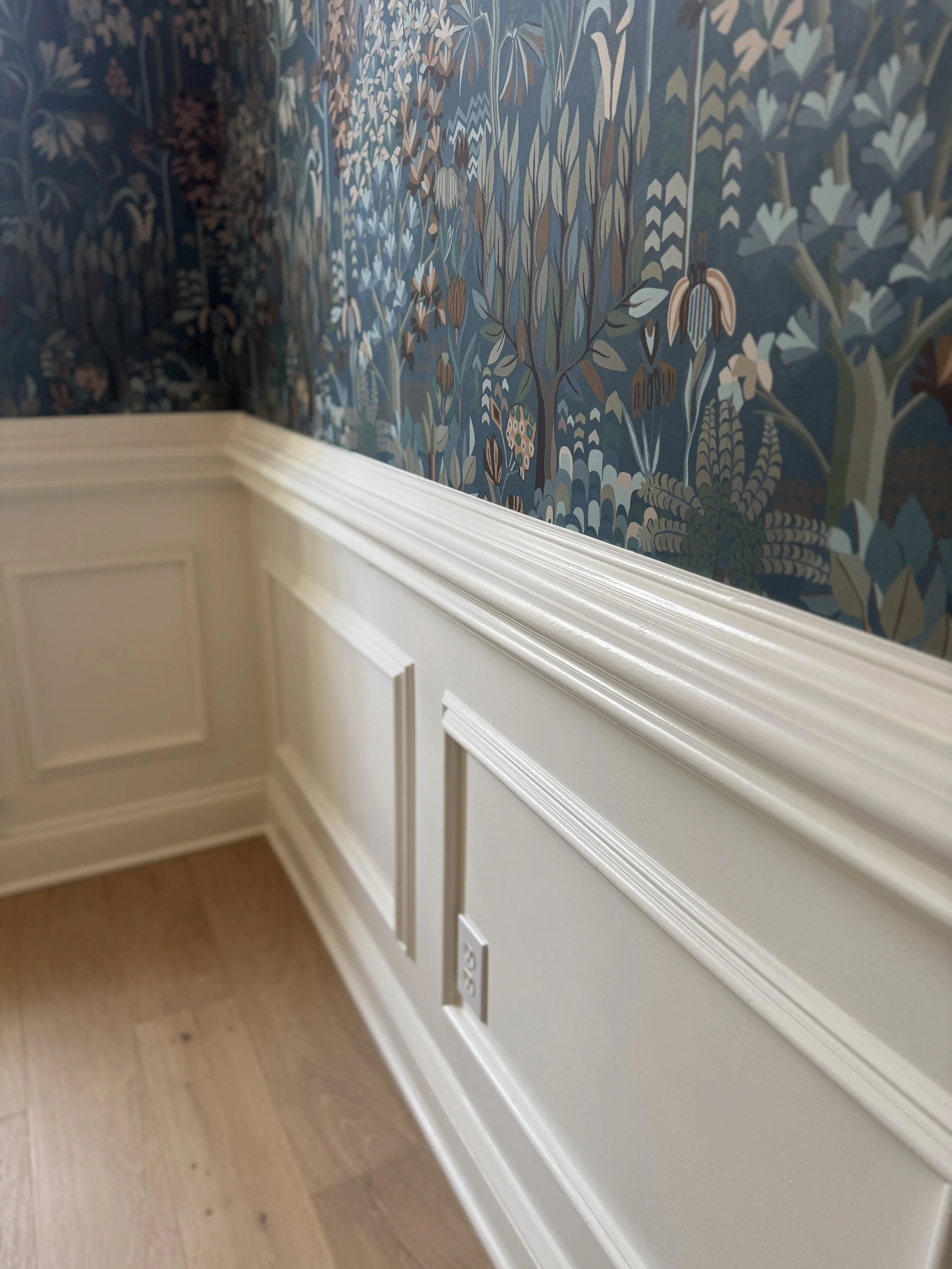 Close-up of a wall with floral patterned wallpaper and white wainscoting with a built-in electrical outlet, hardwood floor.
