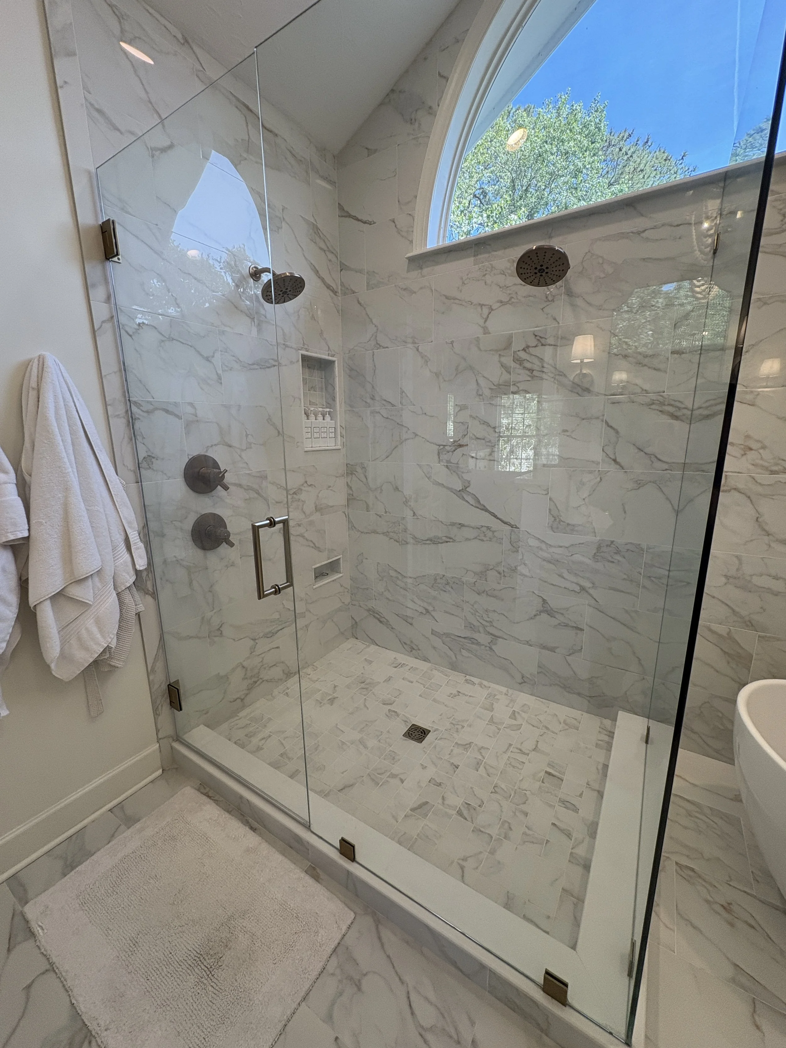 Finished walk-in shower featuring a frameless glass enclosure, oversized marble-look wall tile, and a custom tiled shower pan designed for a seamless, open-concept appearance. Natural light from the dramatic arched window enhances the bright, spa-ins