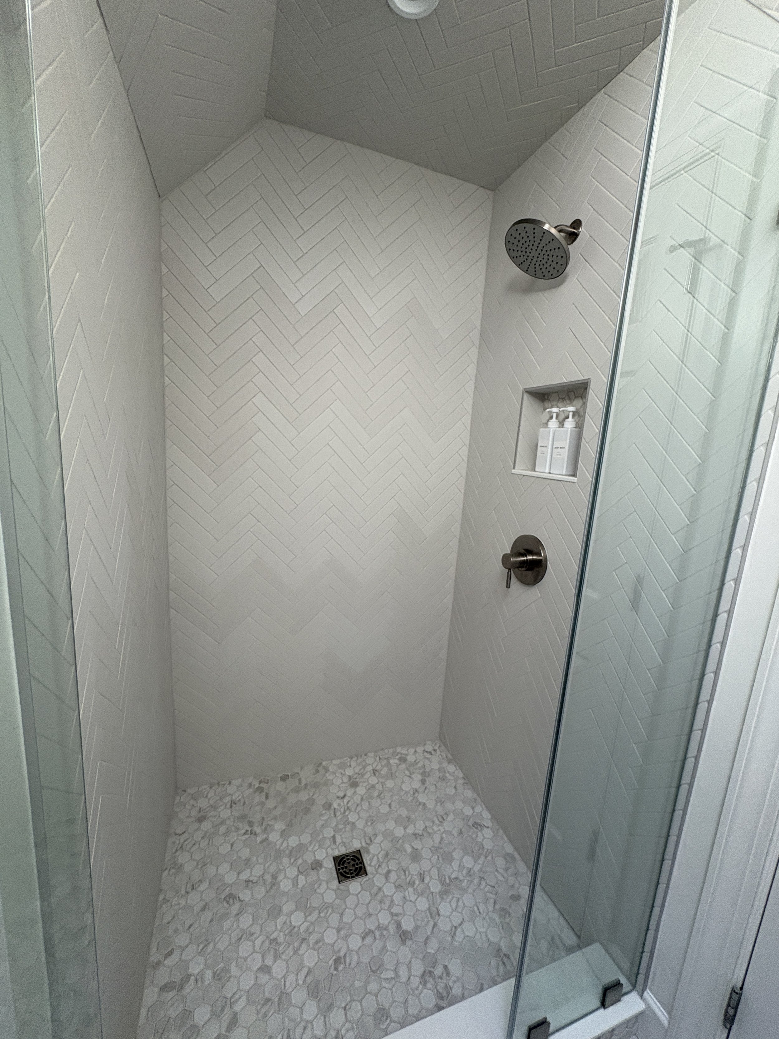 Finished custom walk-in shower featuring a frameless glass enclosure, textured herringbone-pattern wall tile, and a coordinating mosaic tile shower floor that adds visual interest and durability. The clean lines, recessed shower niche, and neutral co