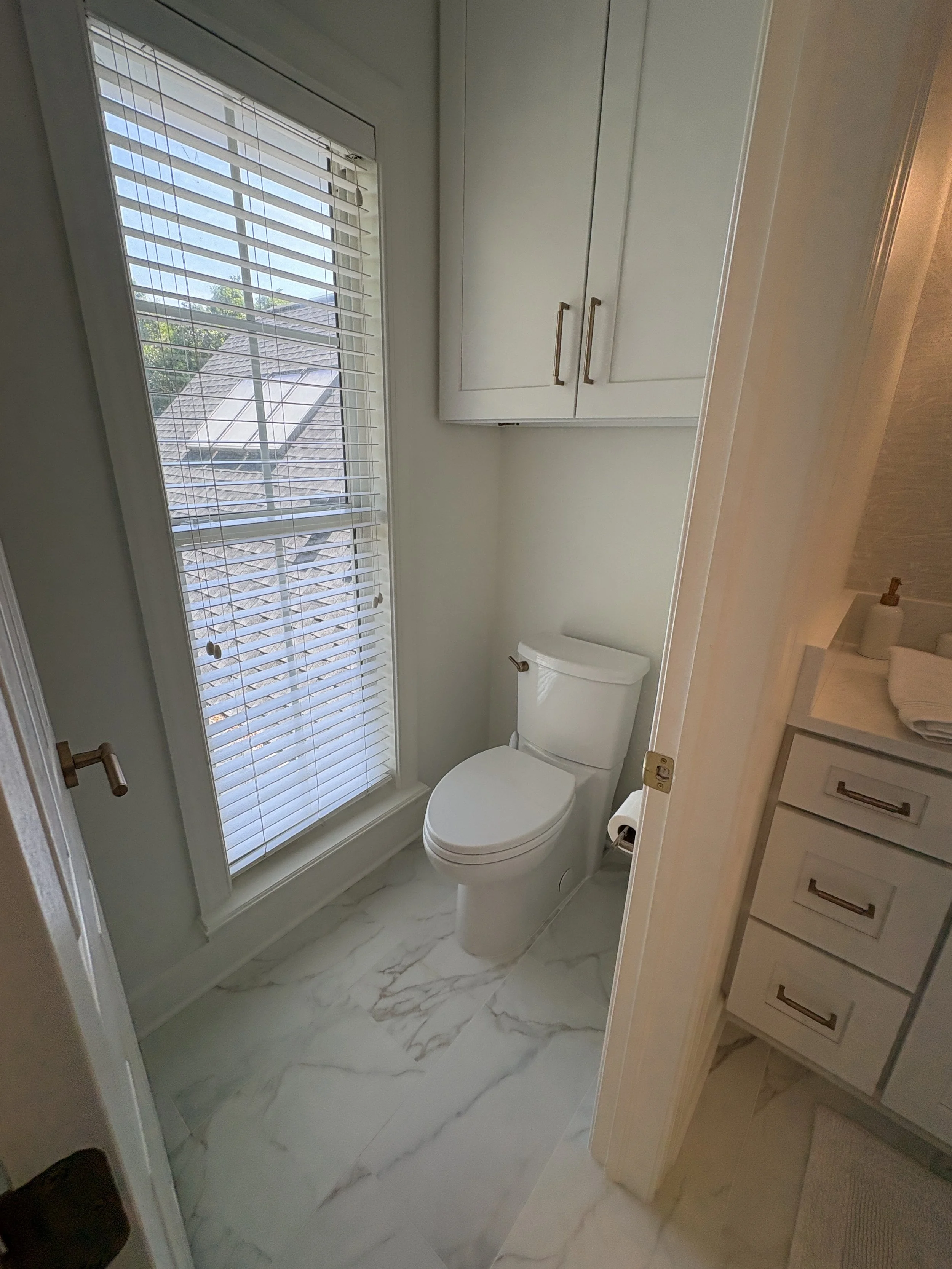 Finished private water closet featuring a modern toilet, custom upper storage cabinetry, and large-format marble-look porcelain tile flooring that seamlessly matches the adjoining primary bathroom. A tall window with plantation shutters provides abun