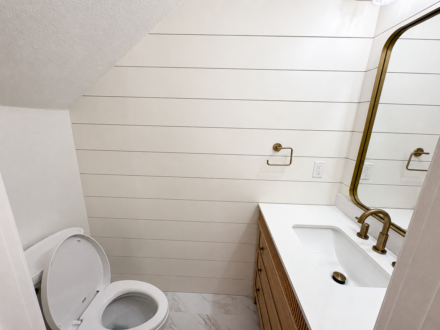 This high-end Jacksonville condo bathroom remodel pairs a custom natural-wood vanity with a sleek white countertop, brushed gold fixtures, shiplap walls, and marble-look tile flooring. Completed by Elite Construction of JAX.