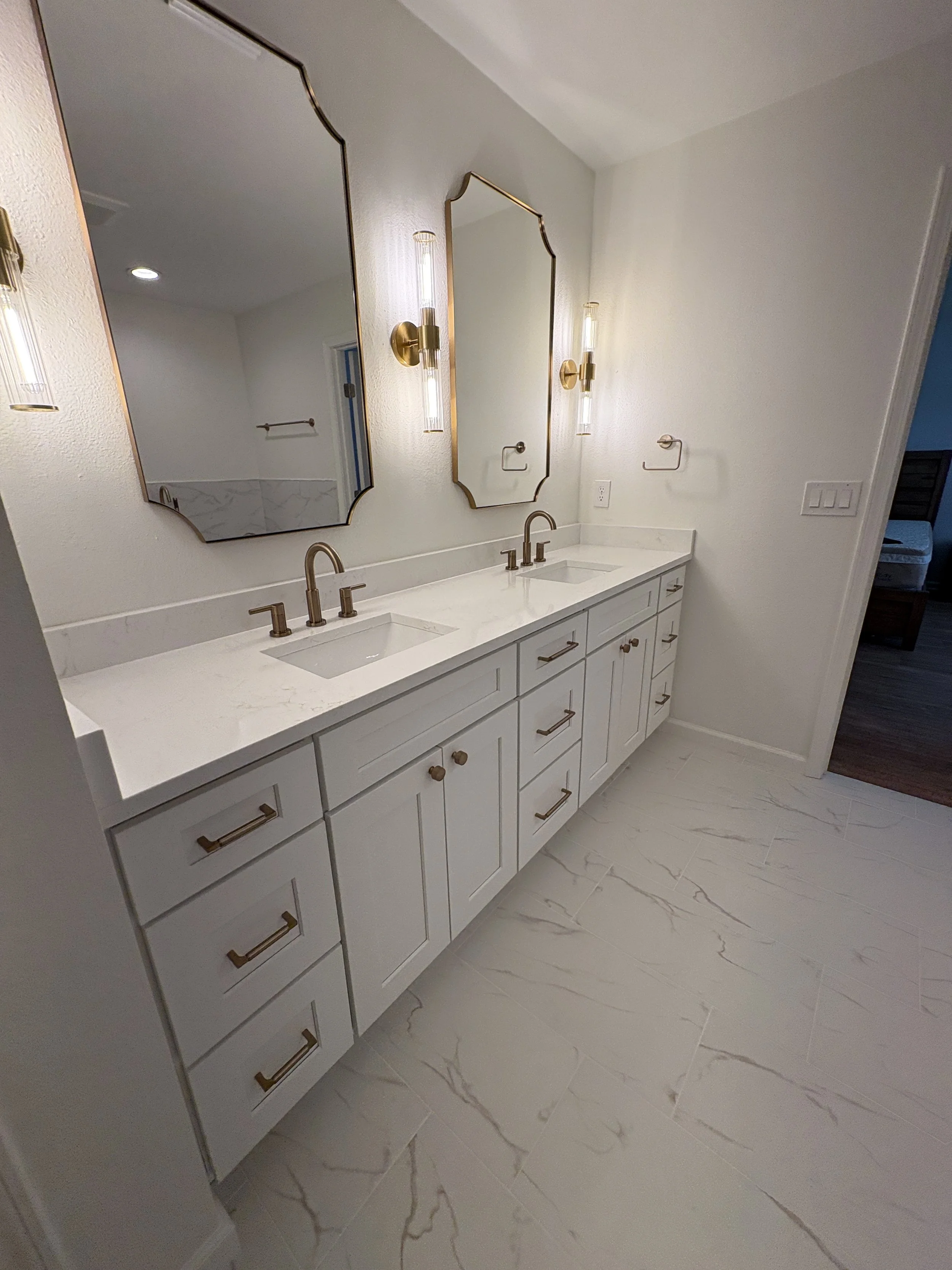 A custom double vanity with extensive storage, a sleek white countertop, decorative mirrors, and brushed gold fixtures brings elegance and function to this luxury Jacksonville condo bathroom remodel by Elite Construction of JAX.