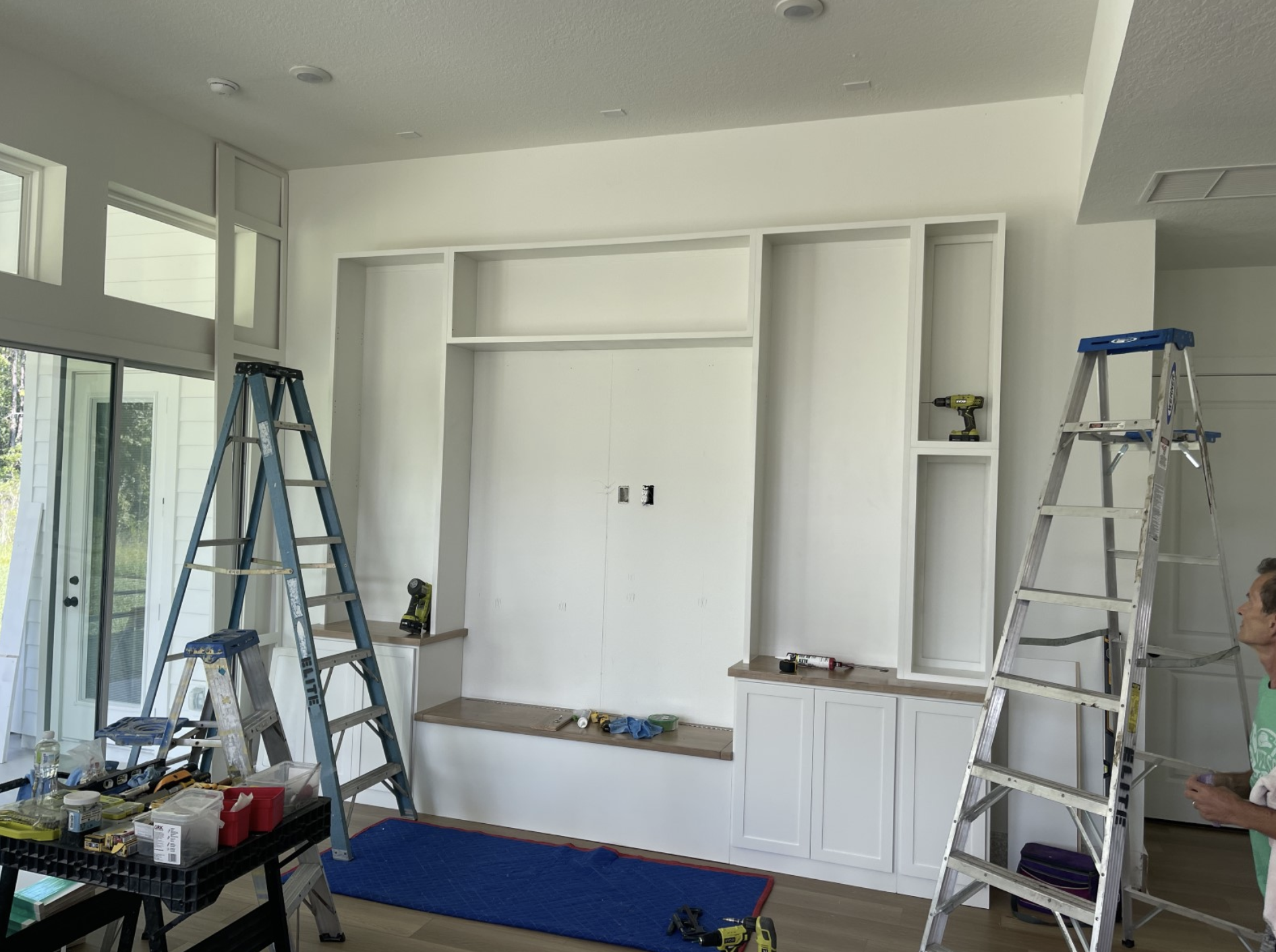 Living room with built-in shelving unit under construction, two ladders, tools, and a worker; large window and glass door to the left.