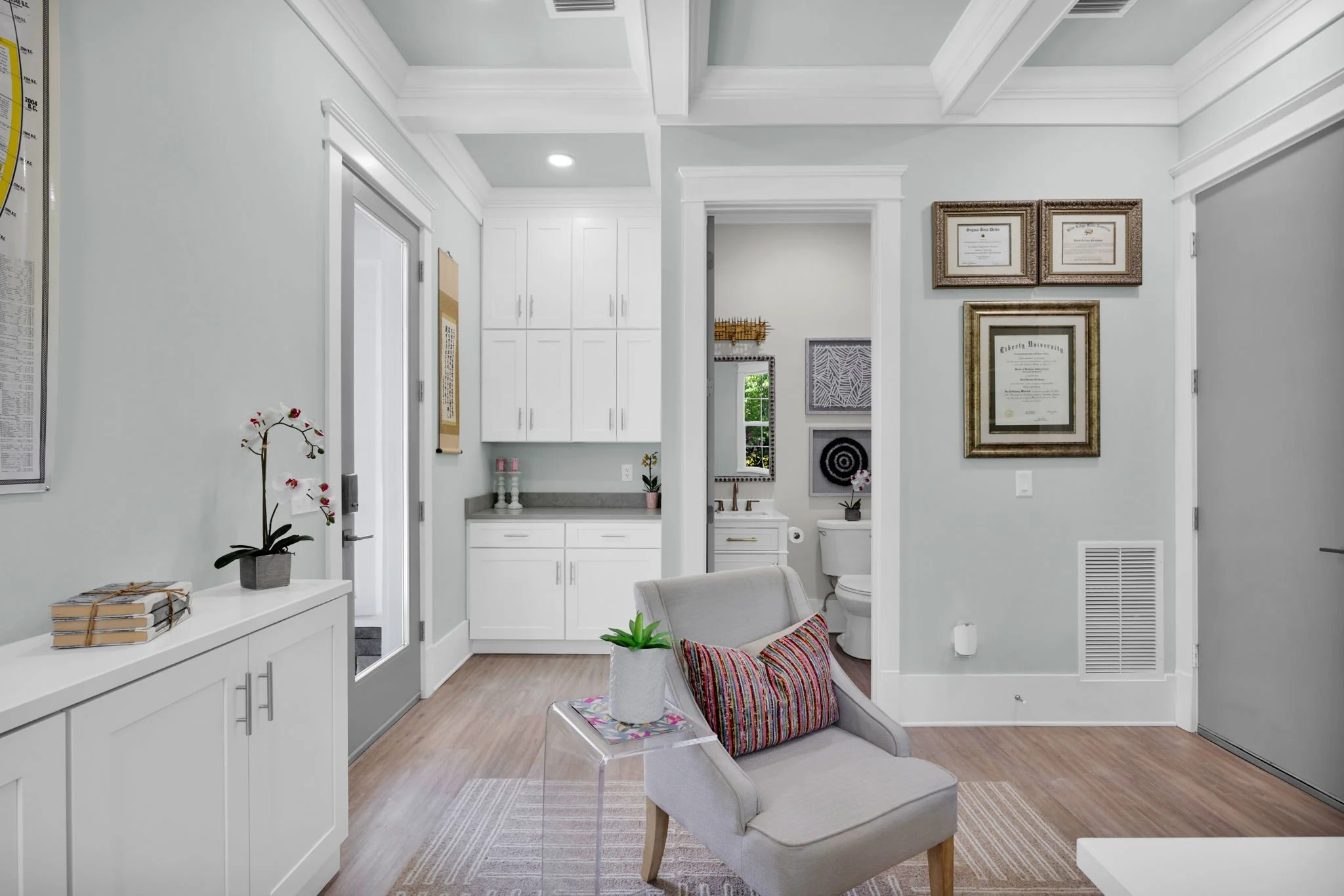 A cozy waiting area with a white armchair, a colorful striped pillow, and a small white side table with a potted plant. Behind, doors lead to a small bathroom, and walls are decorated with framed certificates and artwork. Light-colored wooden floorin