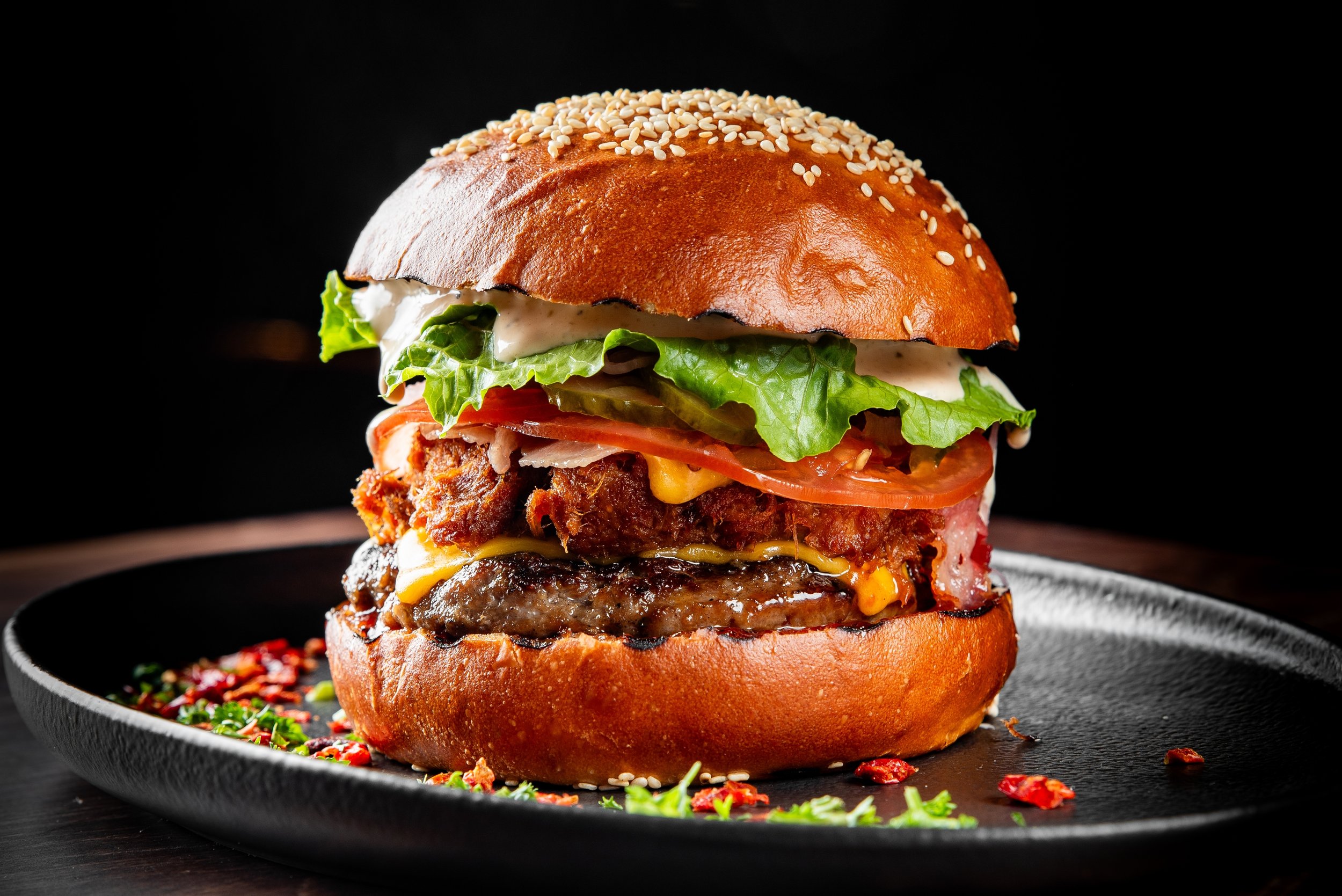 A close-up of a large burger with a sesame seed bun, lettuce, tomato, cheese, bacon, and fried chicken patty, on a black plate with scattered herbs and spices, against a black background.