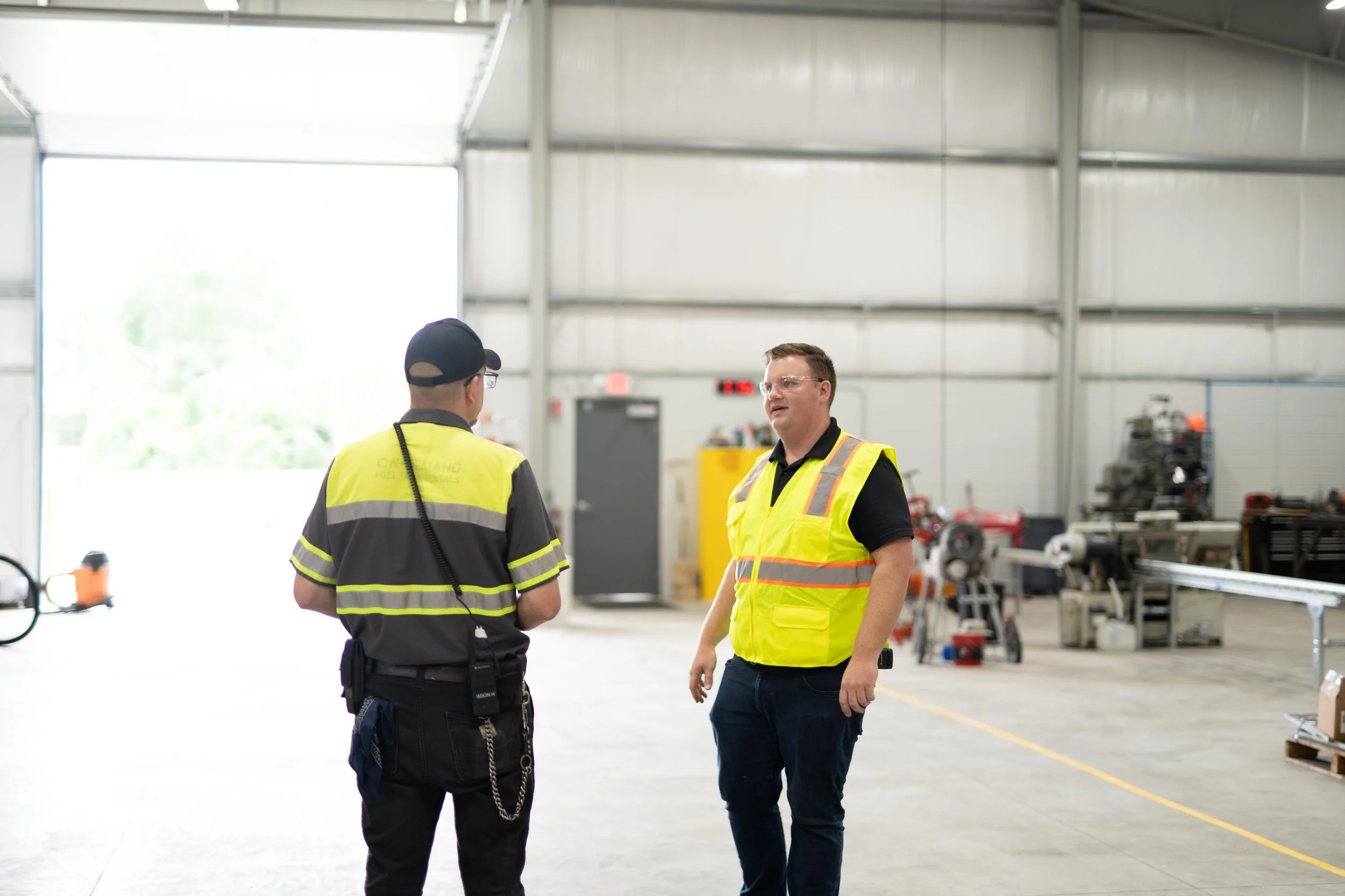 Two workers wearing high-visibility vests talking inside a spacious industrial warehouse with machinery in the background.
