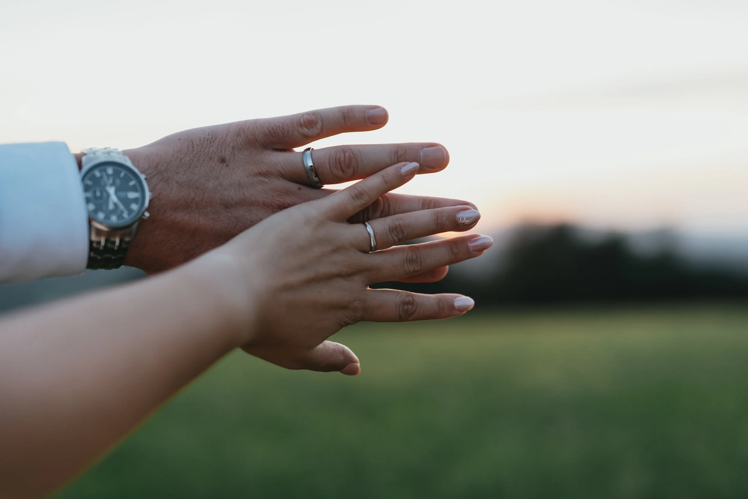 Close-up of couple's hands with wedding bands against a blurred outdoor background at sunset.