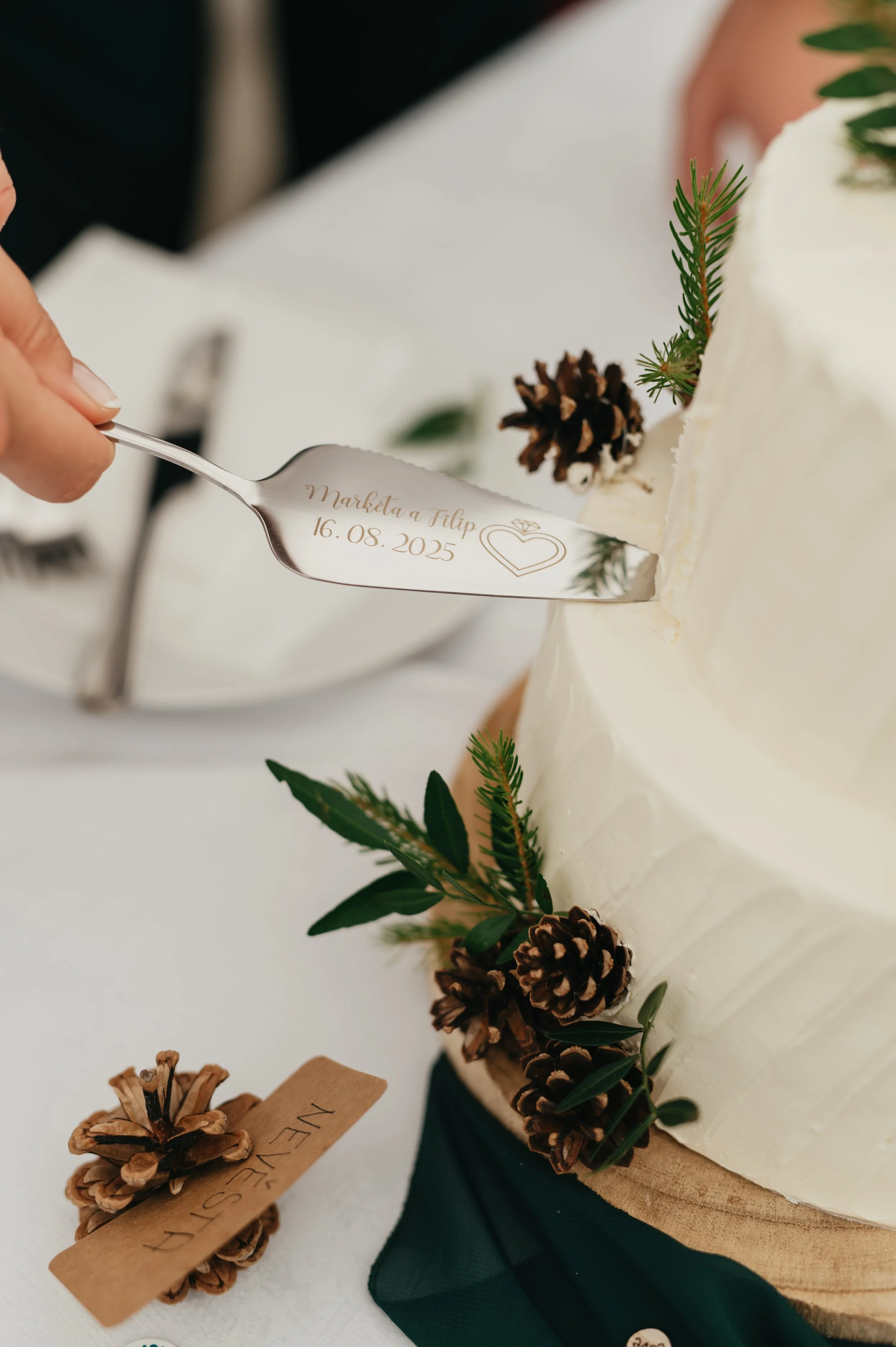 A wedding cake decorated with pinecones, greenery, and a small piece of paper with the word 'NIEST' on it. A hand is carving a message into the cake with a silver cake knife engraved with the names 'Marketa & Filip' and the date '16.08.2025.'
