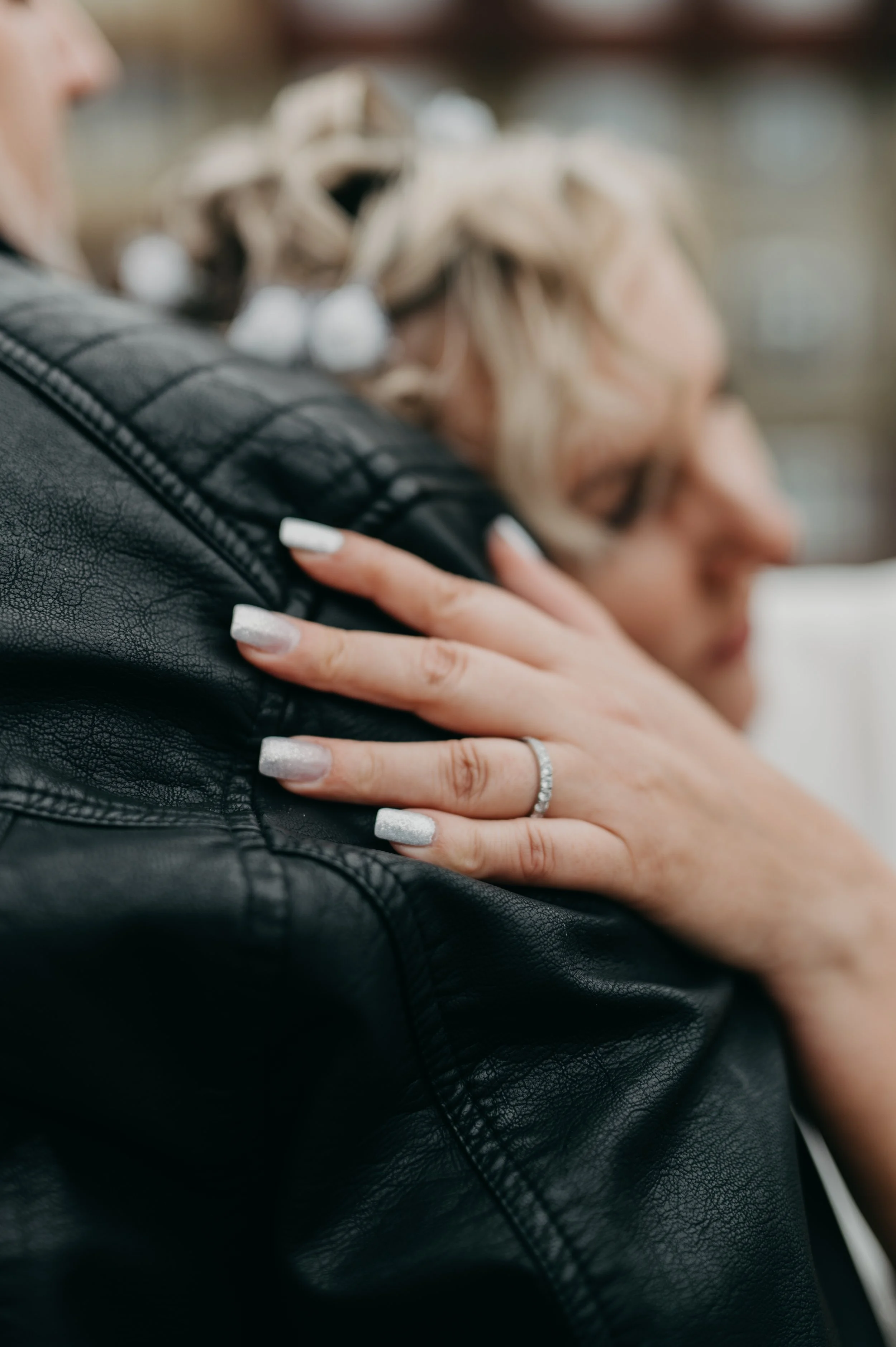 A woman with blonde hair, wearing a ring, resting her head on a man's shoulder, who is wearing a black leather jacket.