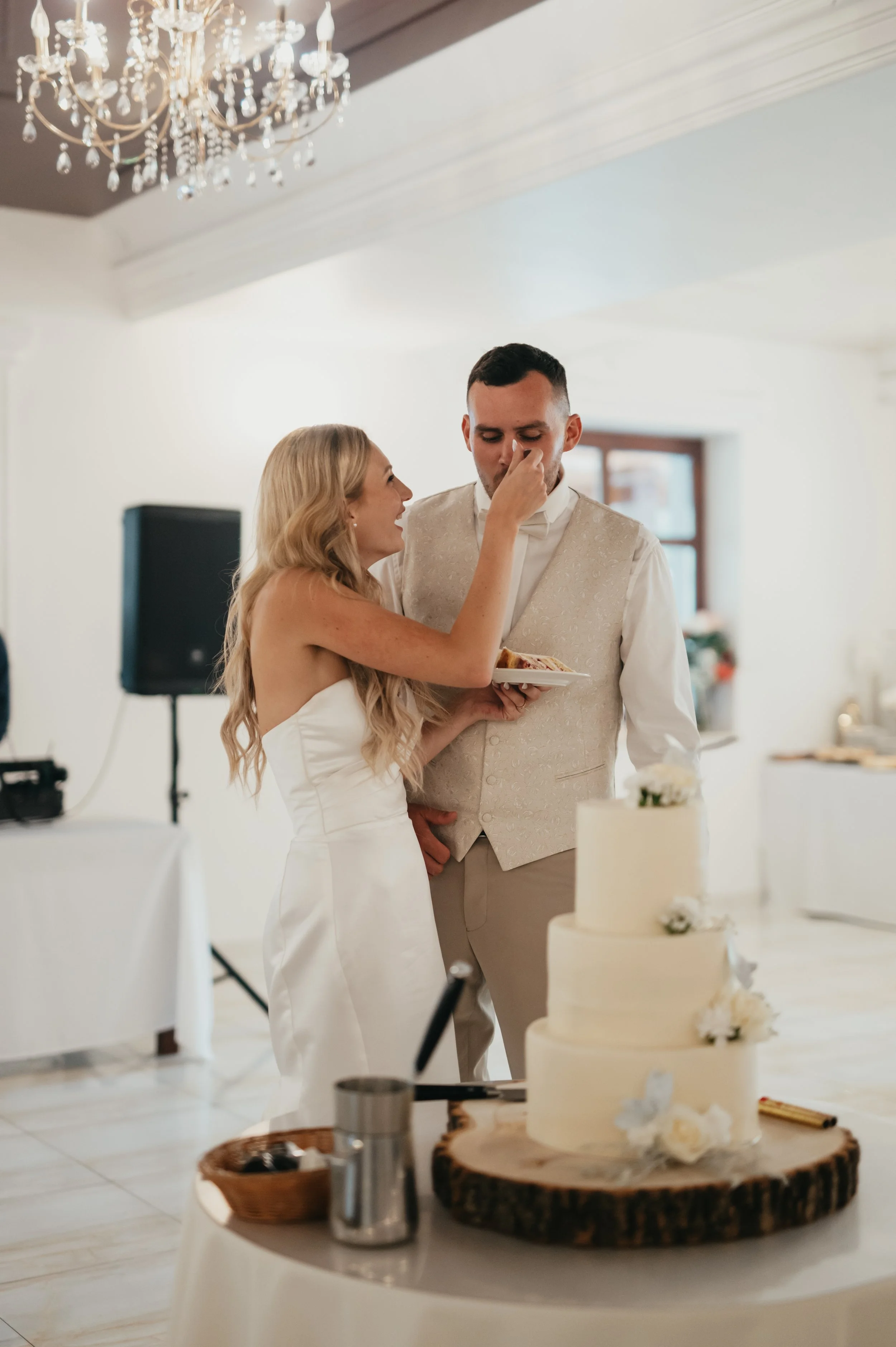 A bride and groom sharing a moment during their wedding reception, with the bride feeding the groom a piece of cake from a plate, in a decorated indoor venue with a wedding cake on the table in front.