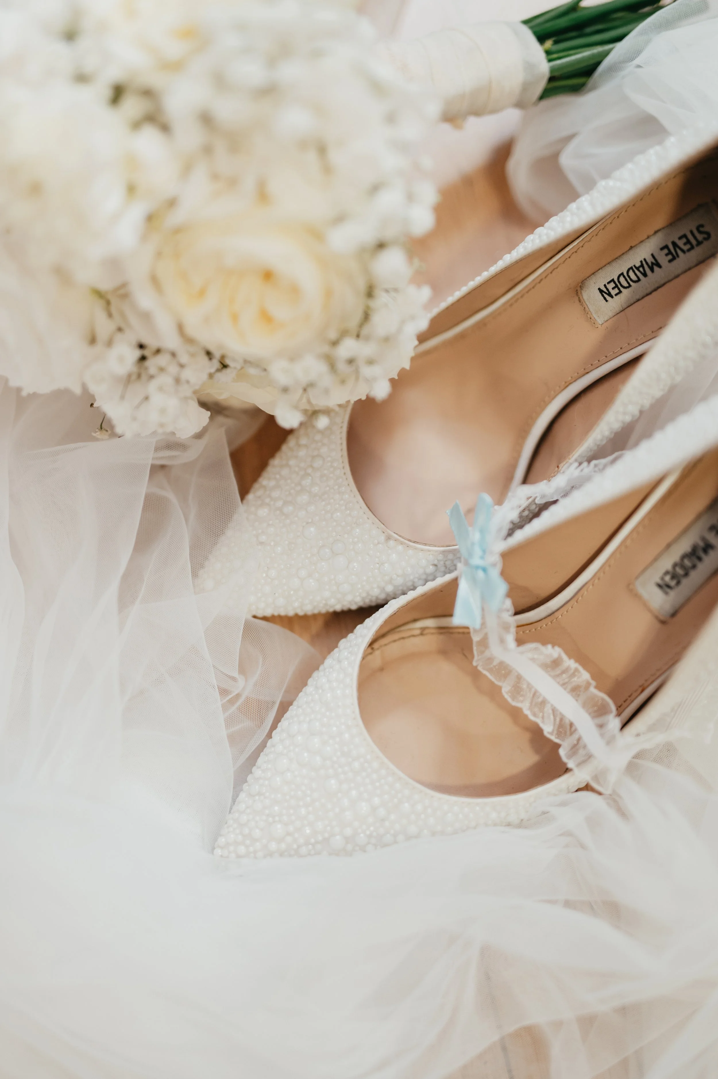 Pár bílých svatebních bot zdobených perlami, umístěných na tylové látce, se svatební kyticí poblíž.
