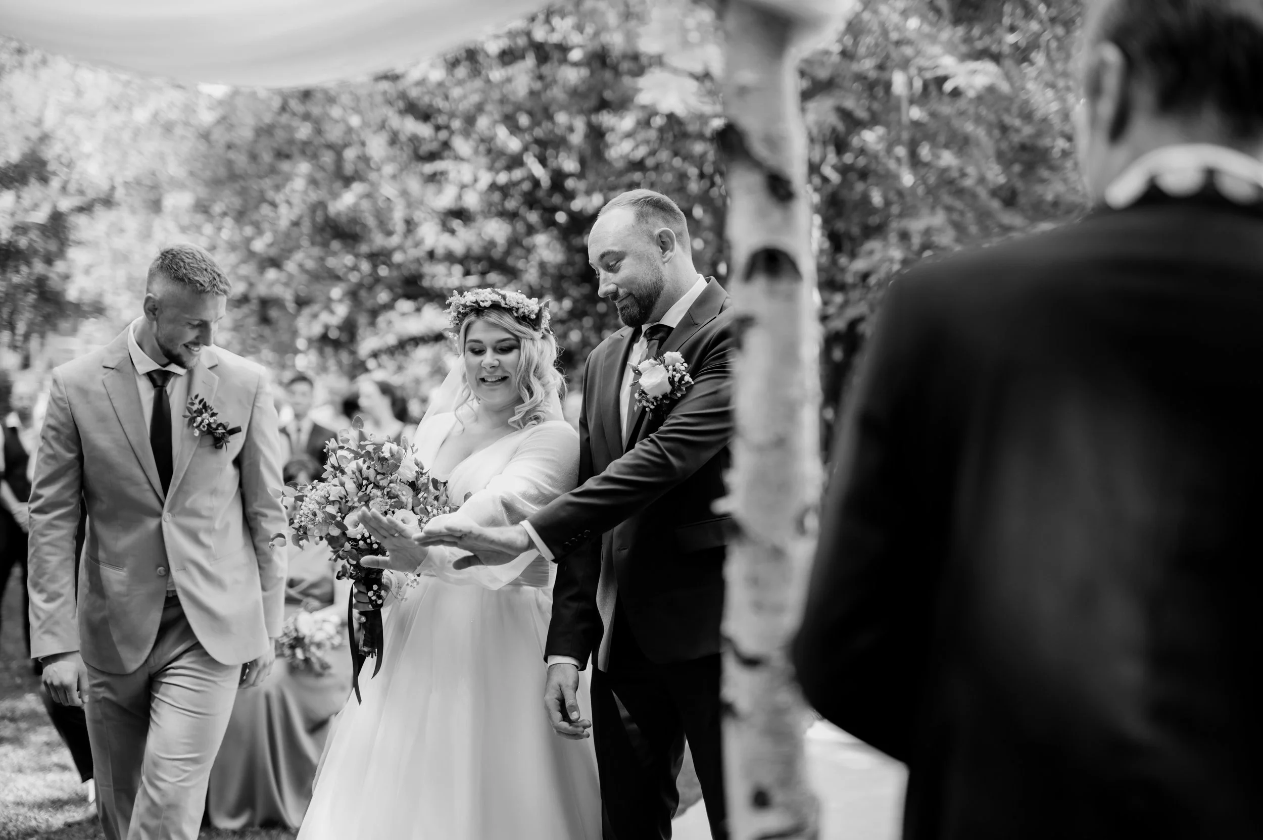 A black and white photo of a wedding ceremony outdoors. The bride and groom are holding hands, surrounded by groomsmen and a guest.