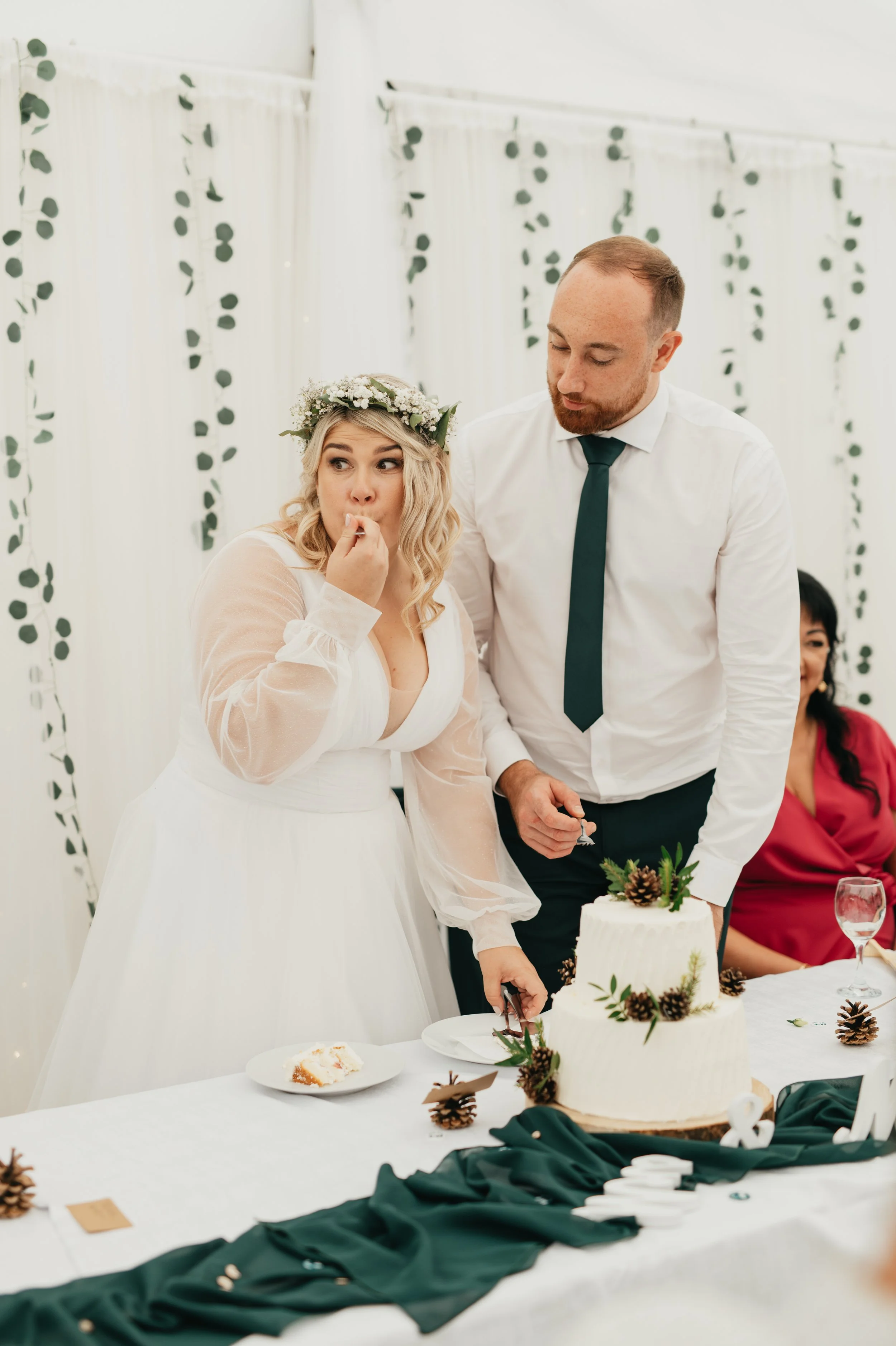 A bride and groom cut a wedding cake at their reception, with the bride wearing a floral crown and the groom in a white shirt and dark tie, while a woman in a red dress watches in the background.