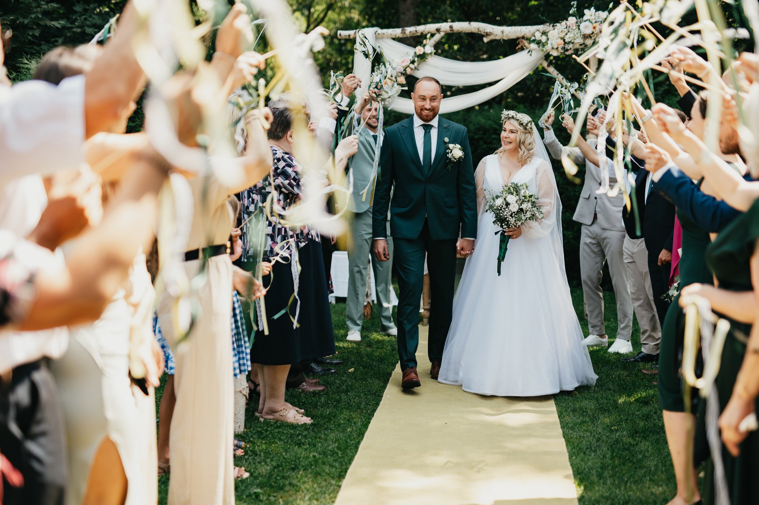 A newlywed couple is walking down the aisle at an outdoor wedding ceremony, holding hands and smiling. The bride, wearing a white wedding gown and a floral crown, holds a bouquet of flowers. The groom, dressed in a dark suit, is smiling beside her. G