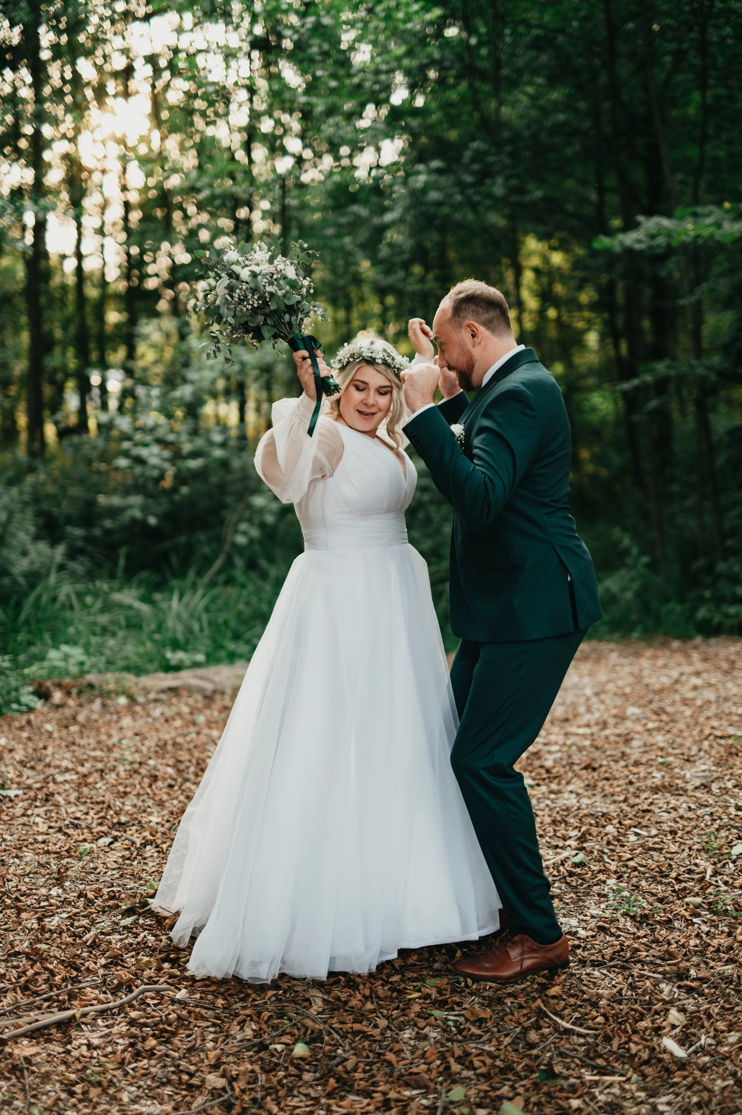 A happy bride and groom dancing outdoors in a wooded area, with the bride holding a bouquet of flowers and wearing a white wedding dress, while the groom is in a dark suit and brown shoes.