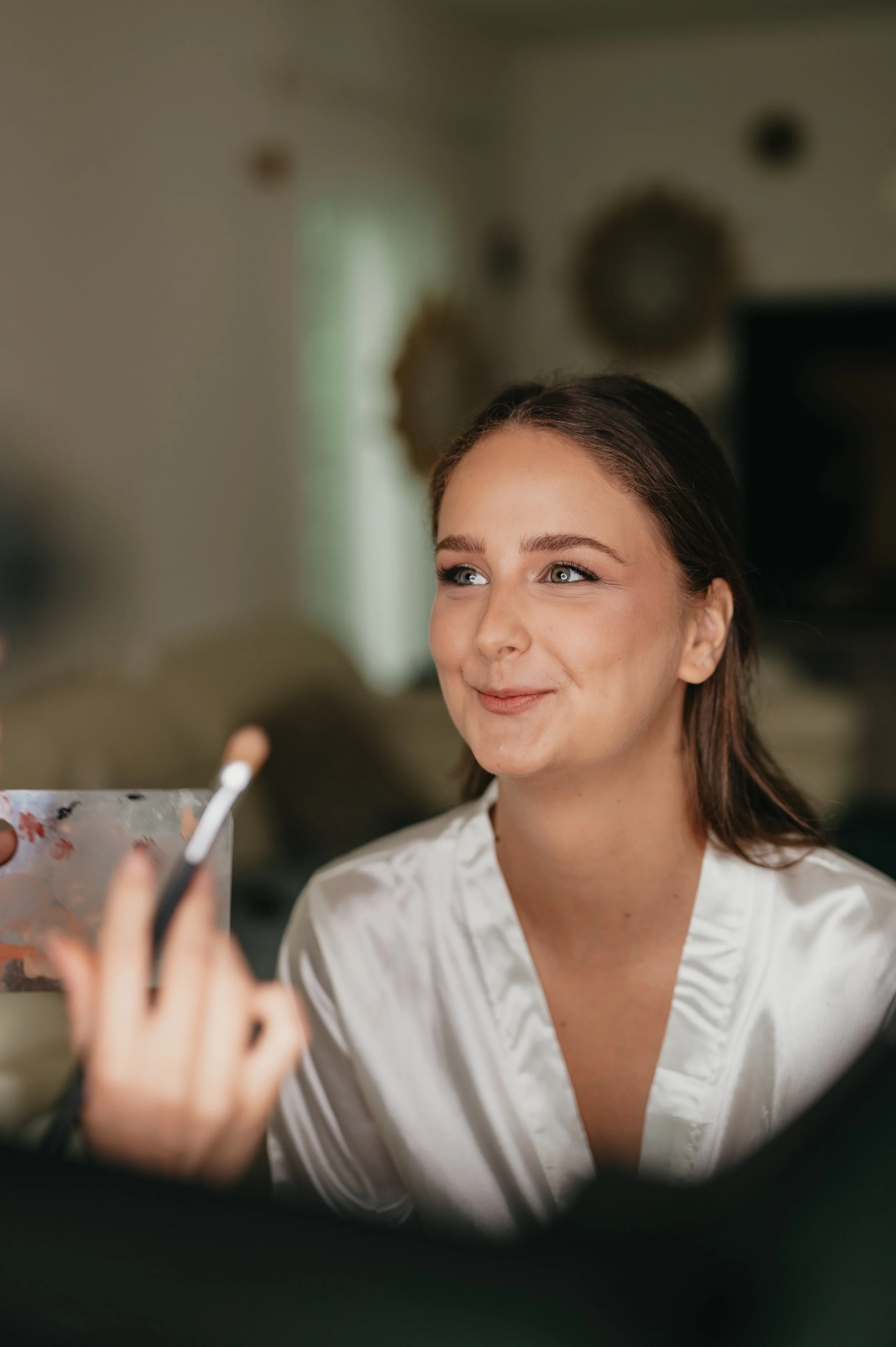Young woman with brown hair smiling softly, wearing a white satin robe, sitting indoors with a blurred background.