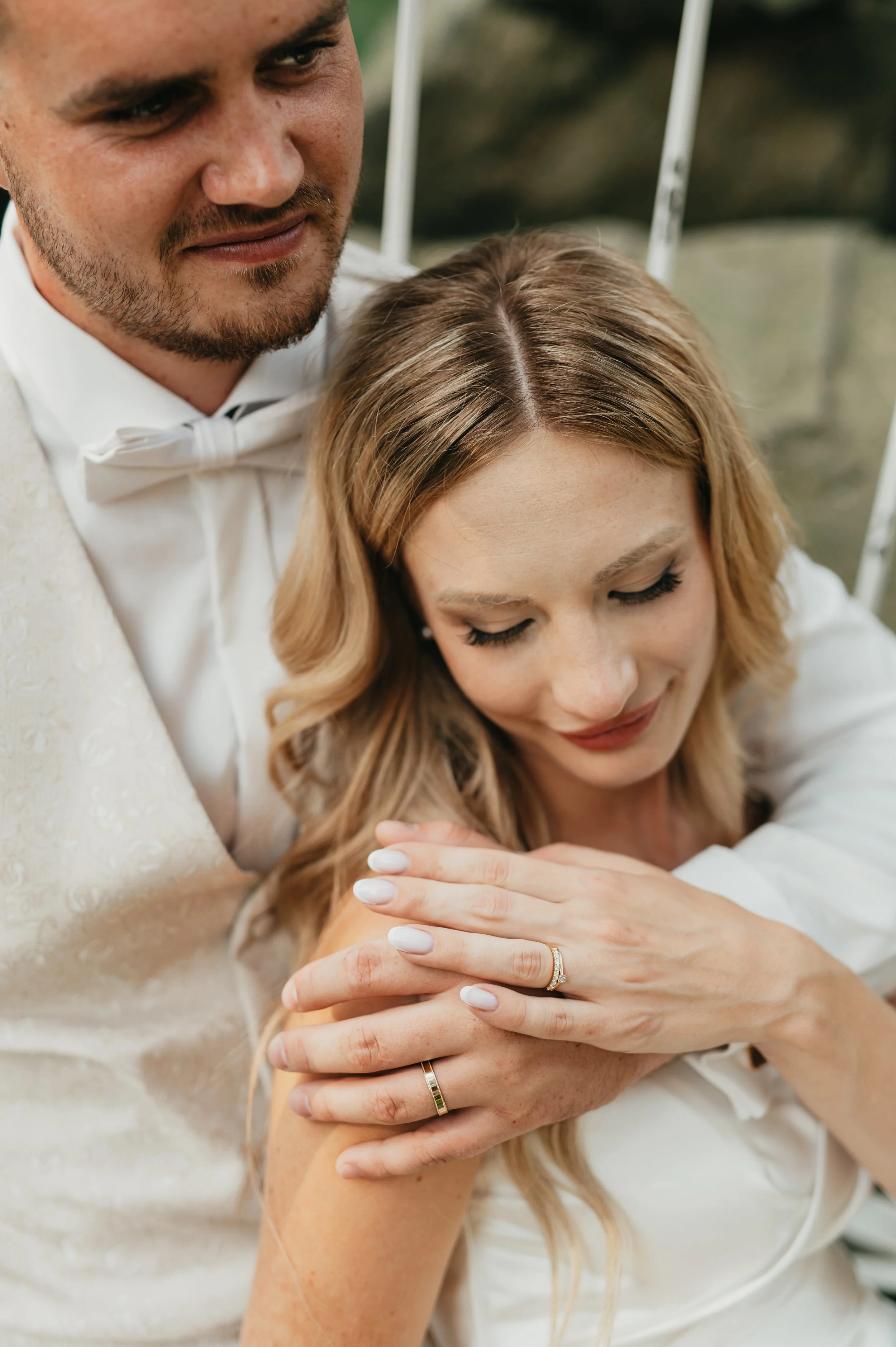 Close-up of a newlywed couple, with the groom gently holding the bride's shoulder. The bride has blonde hair, closed eyes, and an engagement ring on her finger. The groom is wearing a white tuxedo with a bowtie, and the bride's nails are painted whit