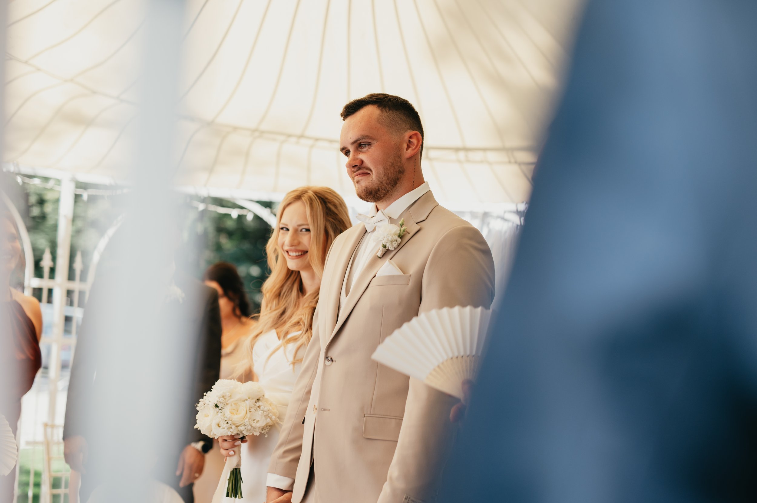 Nevěsta a ženich stojící pod baldachýnem během svatebního obřadu, nevěsta se usmívá a drží kytici bílých květin, zatímco ženich vypadá vážně, oblečený v béžovém smokingu s bílou boutonniérou.