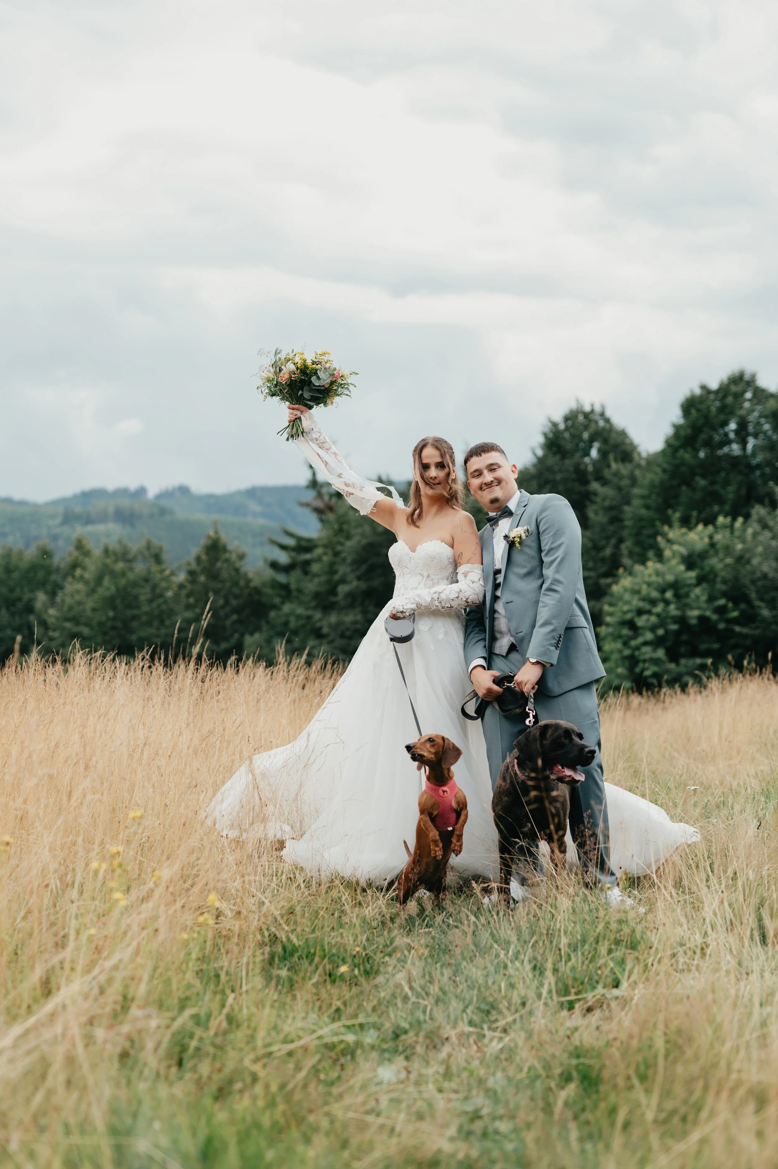 A bride and groom standing in a field with two dogs, one wearing a pink outfit, during their wedding photos.