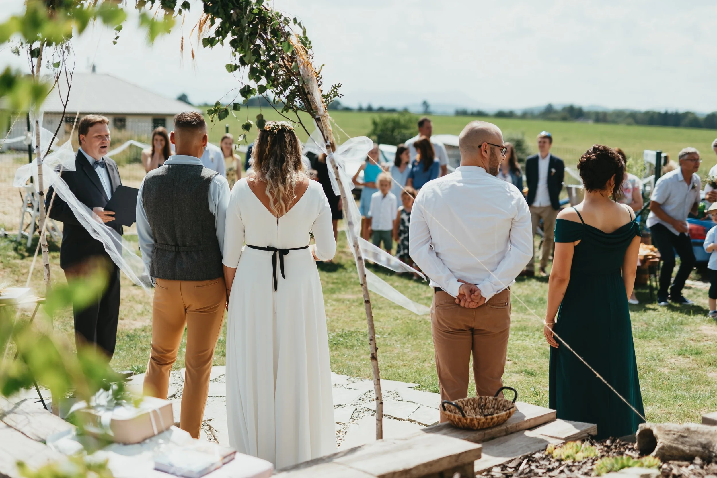 Outdoor wedding ceremony with officiant, bride in white dress, groom, and two guests standing, surrounded by attendees.