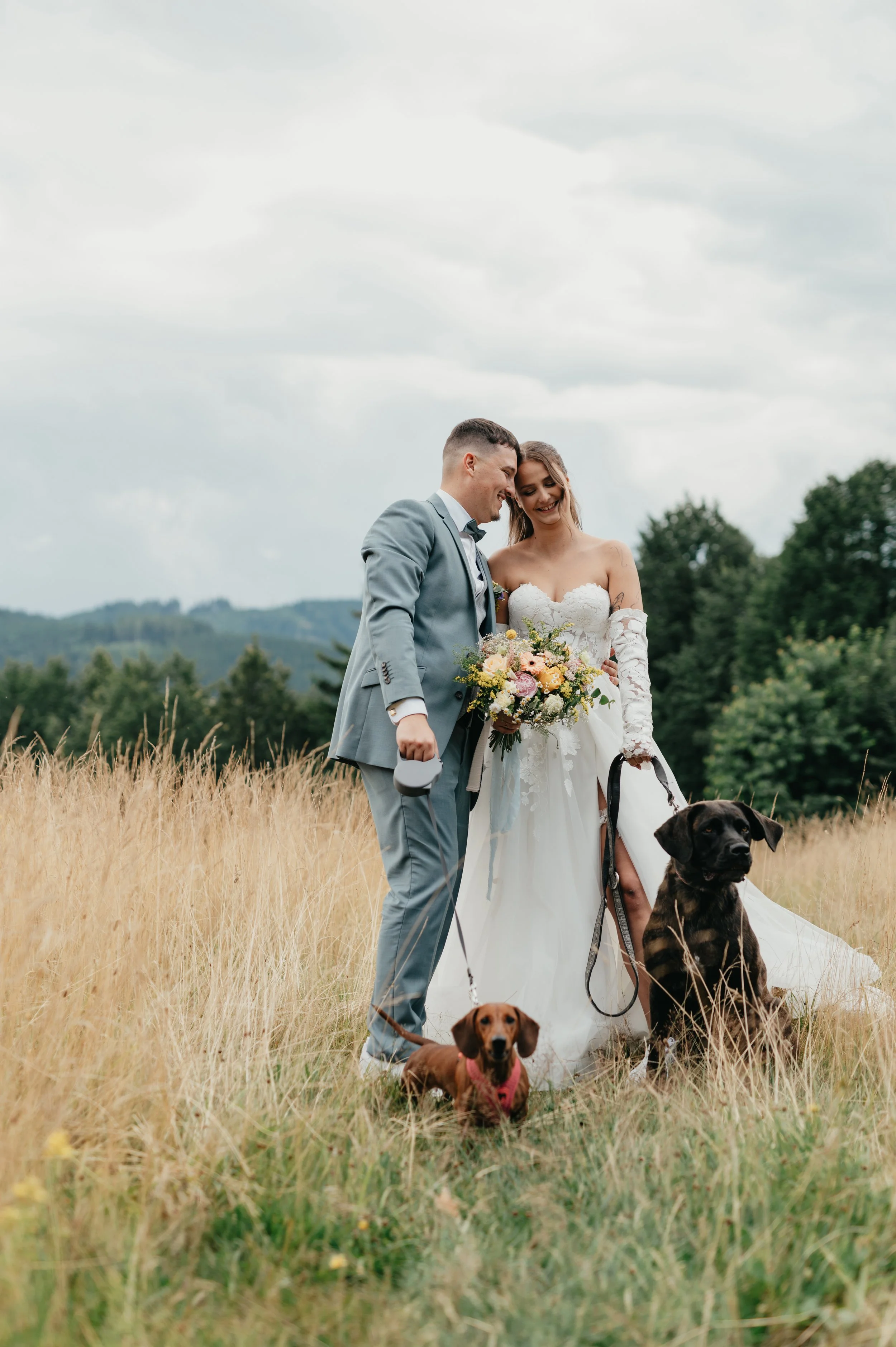Novomanželský pár stojící na poli se svými dvěma psy, ženich v šedém obleku a nevěsta v bílých svatebních šatech držící kytici, nad hlavou zatažená obloha.