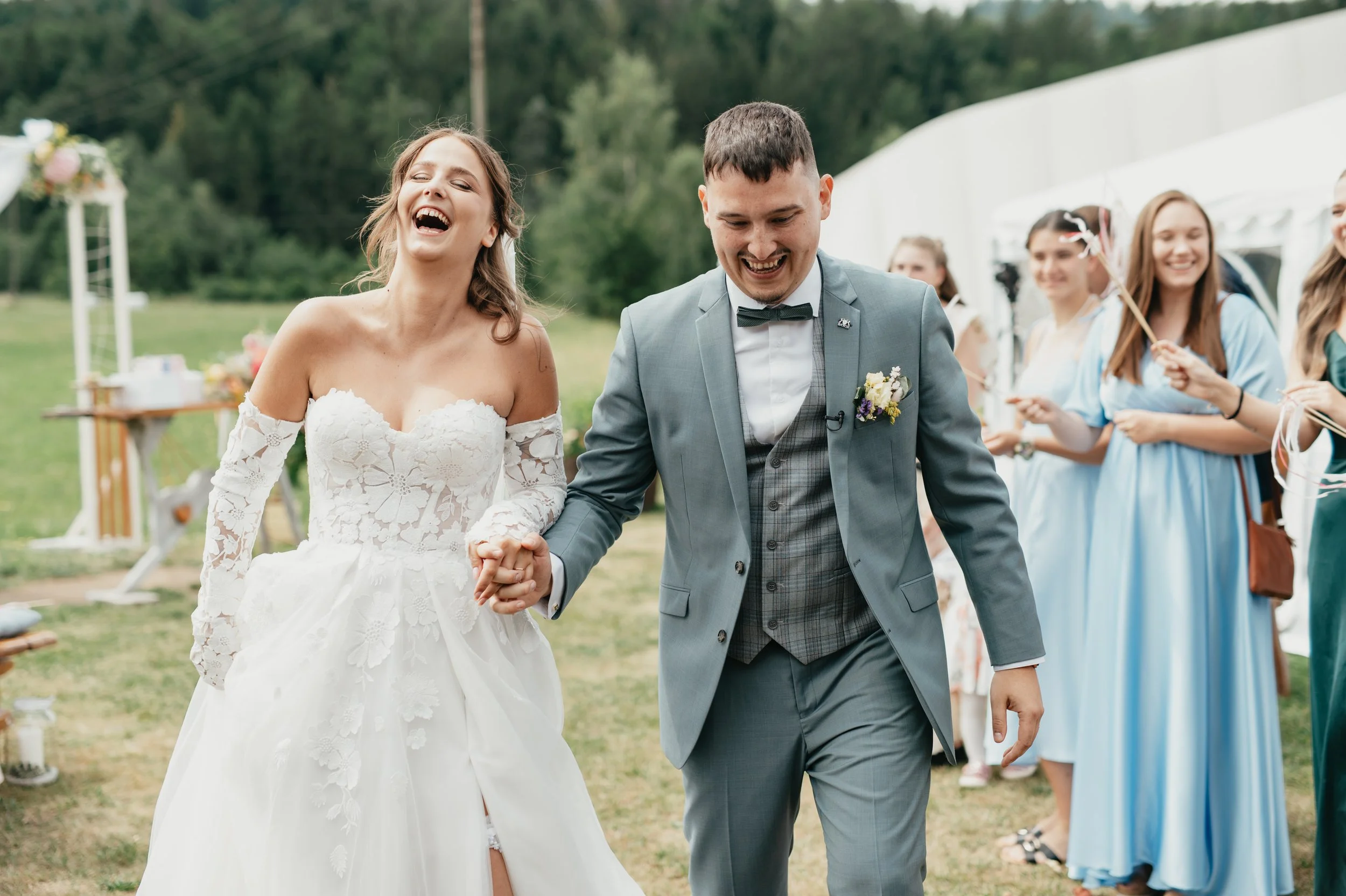 Bride and groom holding hands and smiling at an outdoor wedding reception, with bridesmaids in the background.