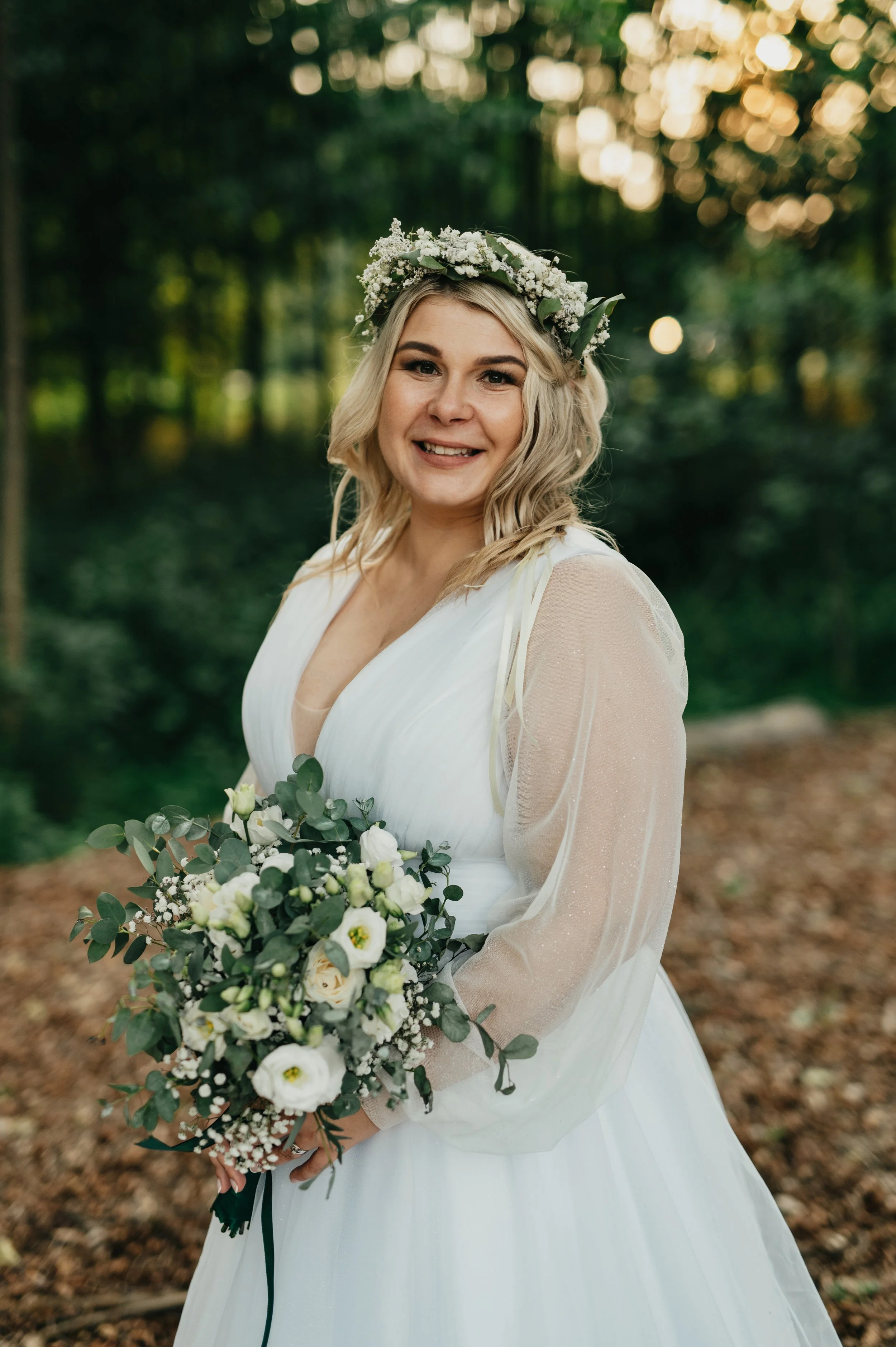 A smiling woman in a white wedding dress holding a bouquet of white and green flowers, wearing a floral crown, standing in a wooded outdoor setting at sunset.