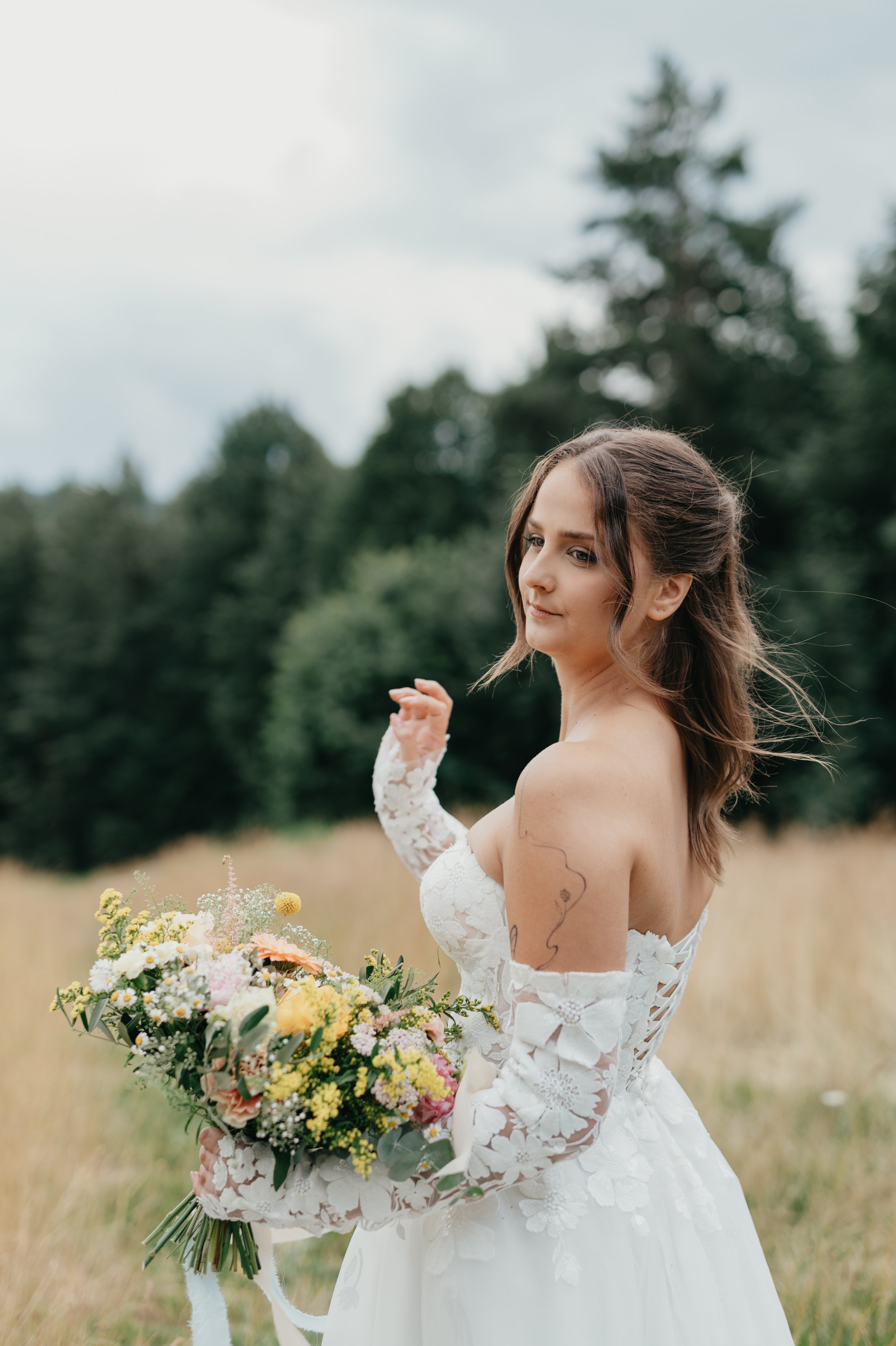 A young woman in a white wedding dress holding a colorful bouquet of flowers outdoors, with trees and a cloudy sky in the background.