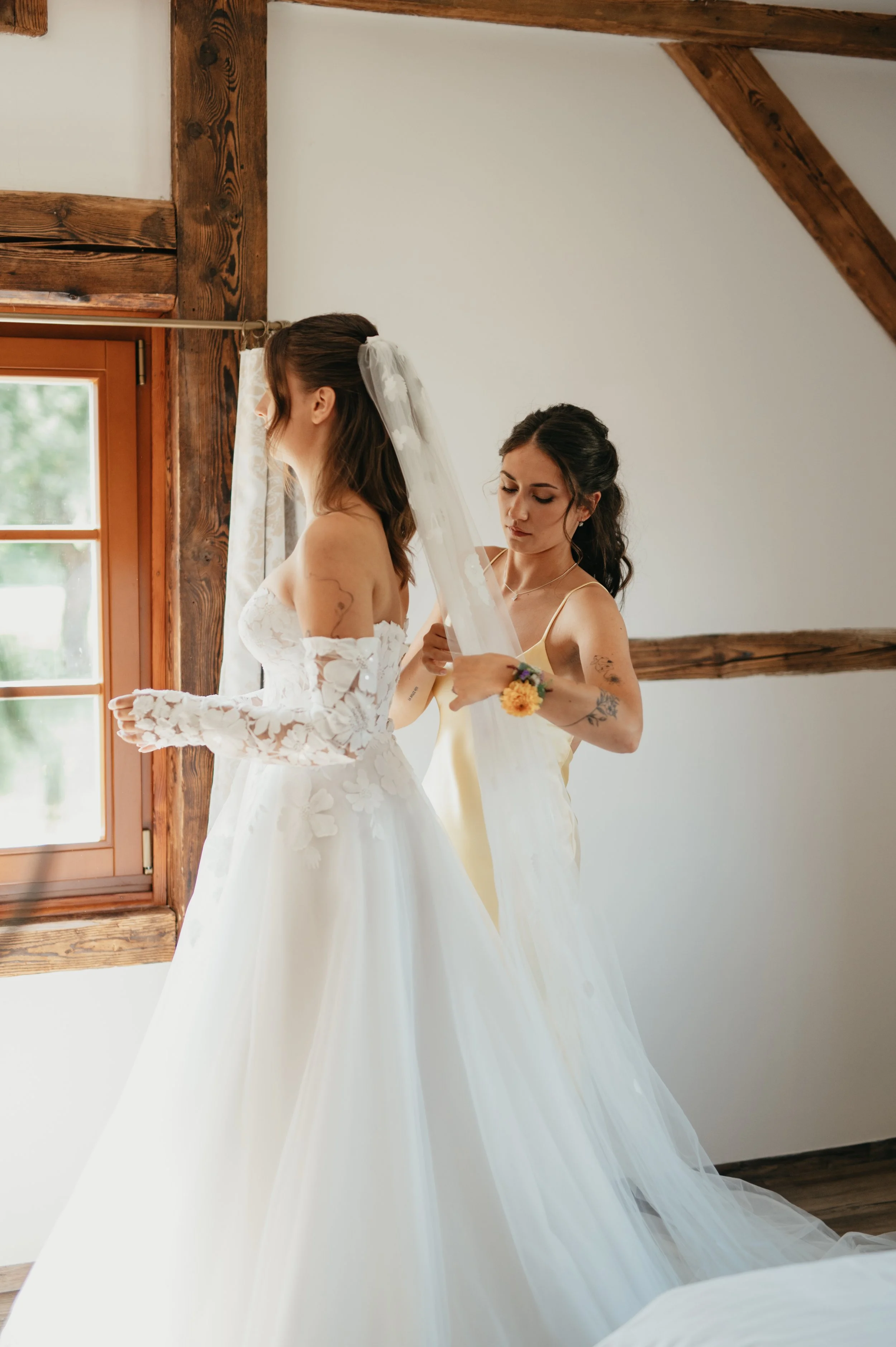 A bride in a white wedding gown with lace details is being assisted by a woman in a yellow dress as she prepares for her wedding.