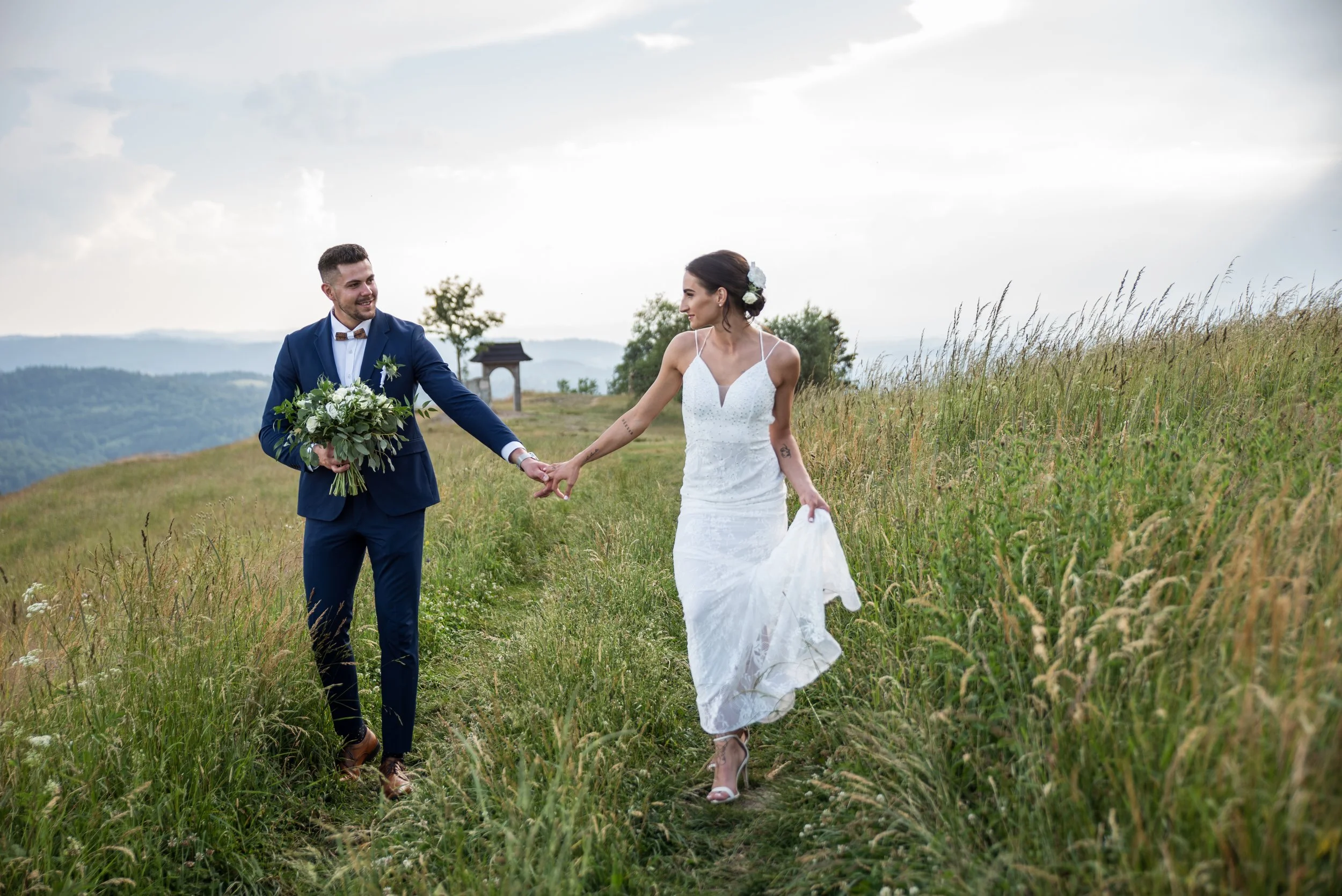 Bride and groom walking on a grassy hill, holding hands, with the groom in a blue suit and the bride in a white wedding dress, carrying a bouquet.