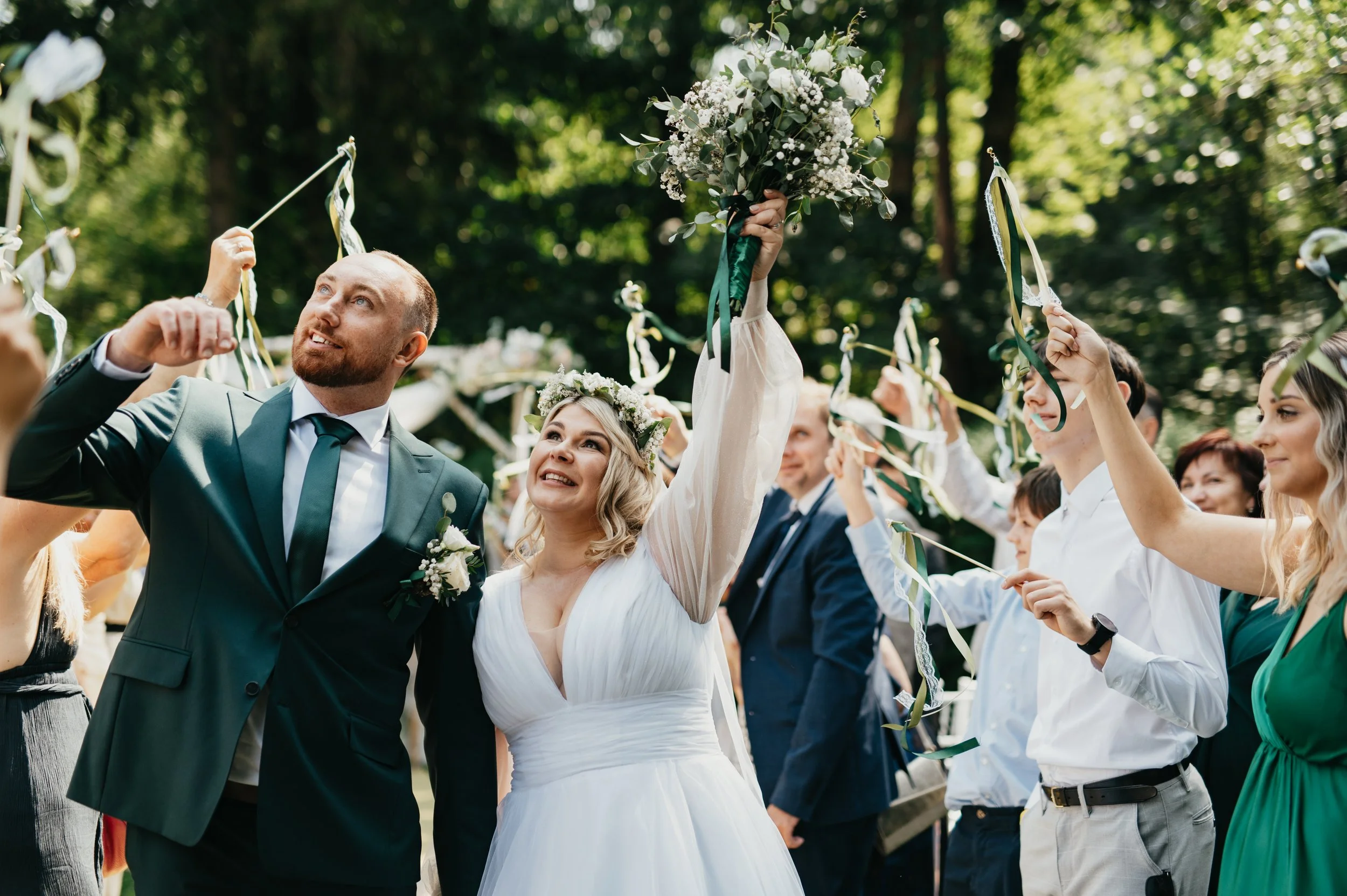 Wedding celebration with bride and groom surrounded by friends outdoors. The bride holds a bouquet and wears a flower crown, while the groom in a suit looks up. Guests hold ribbons and create a festive atmosphere among green trees.