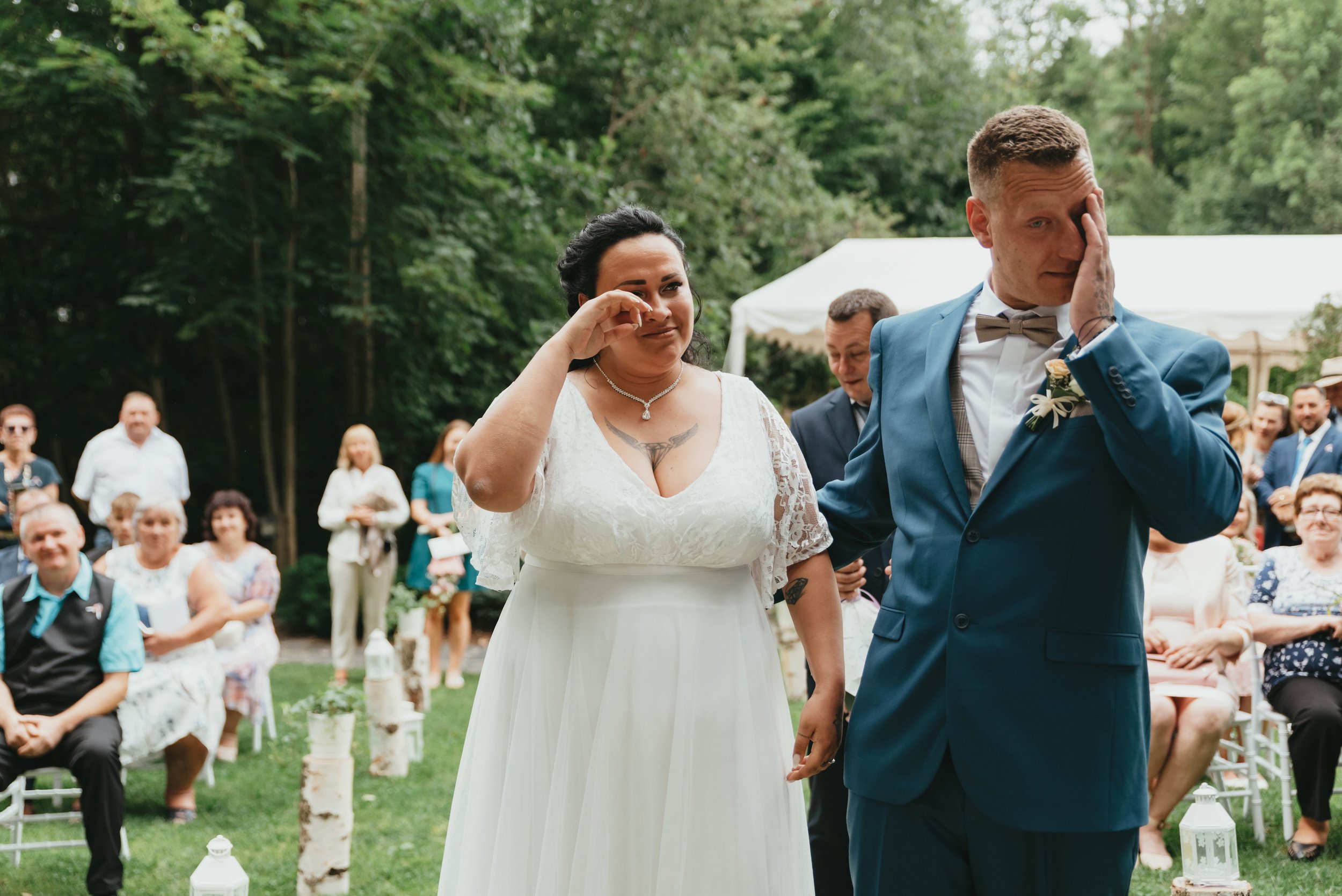 Nevěsta a ženich na svém venkovním svatebním obřadu, nevěsta i ženich si utírají slzy, obklopeni sedícími a stojícími hosty na zeleném trávníku se stromy a bílým stanem v pozadí.