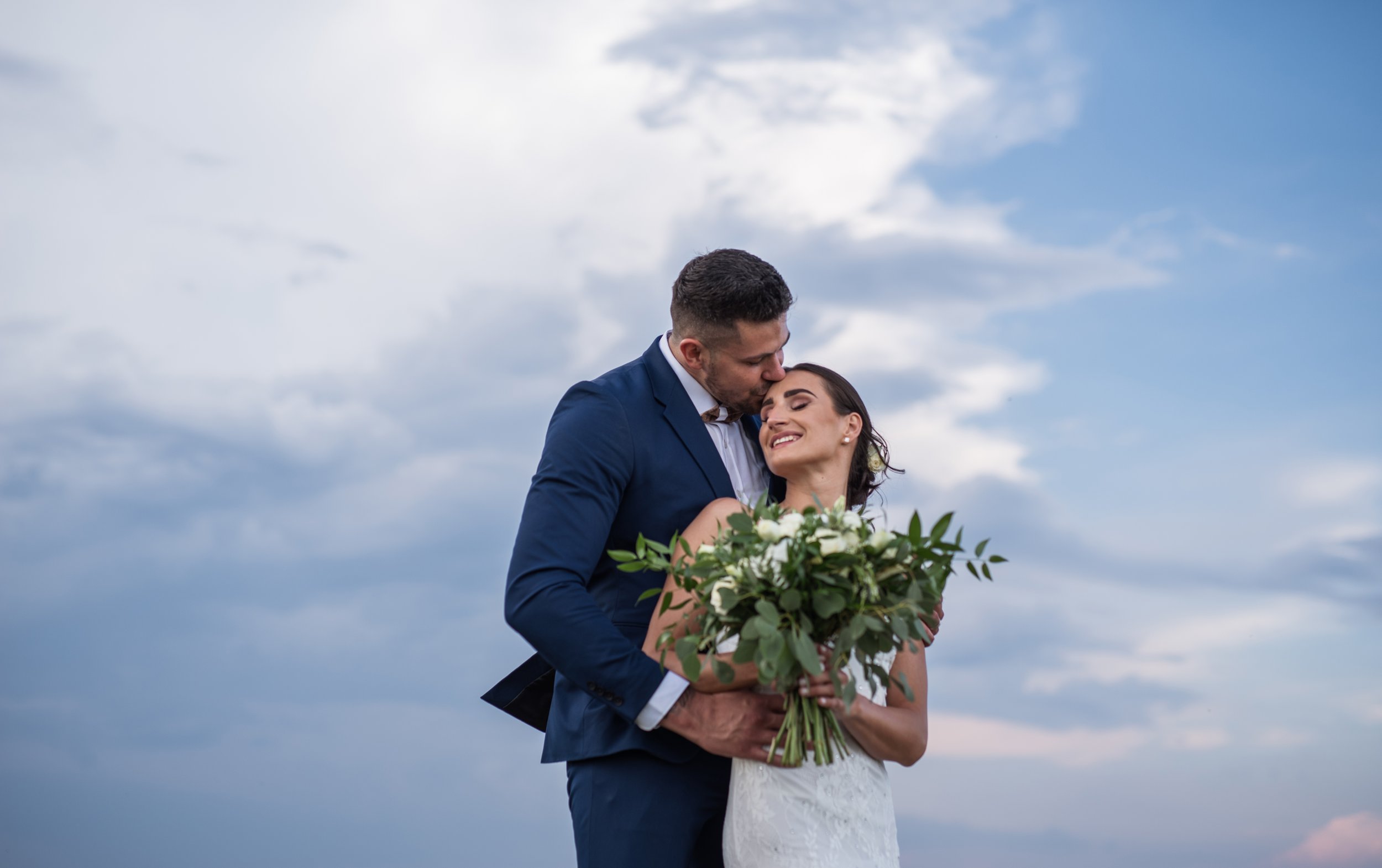 Bride and groom embracing outdoors under a cloudy sky.