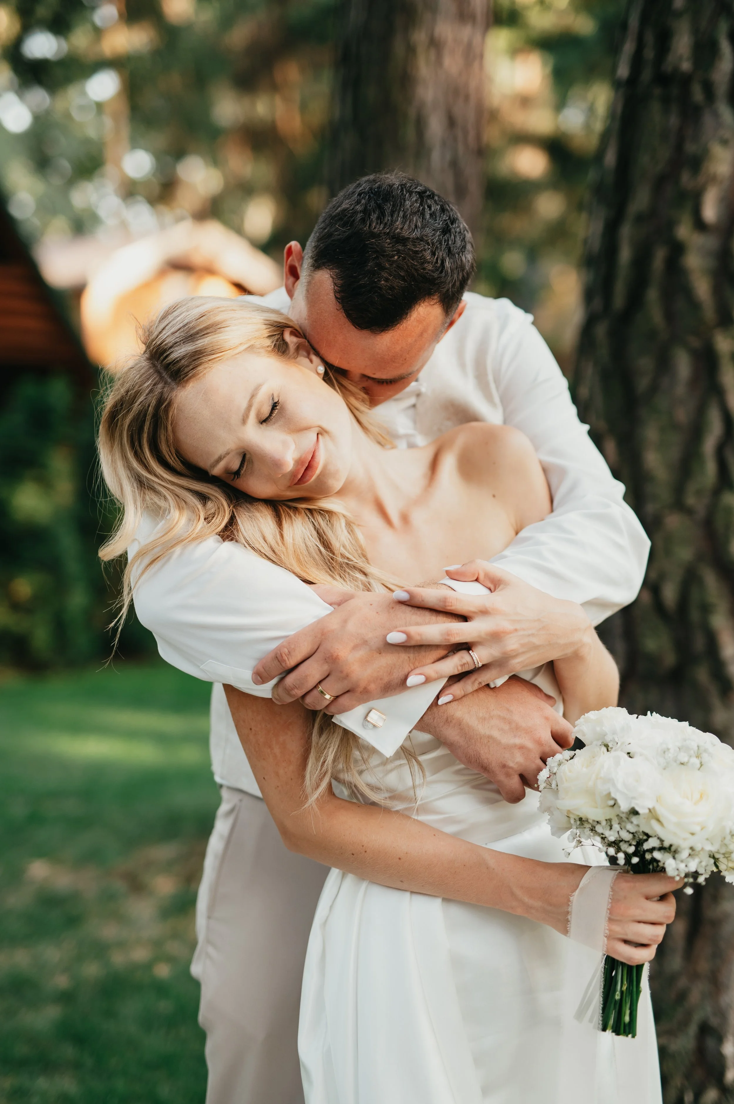 A newlywed couple hugging outdoors with trees in the background, the bride holding a bouquet of white flowers.