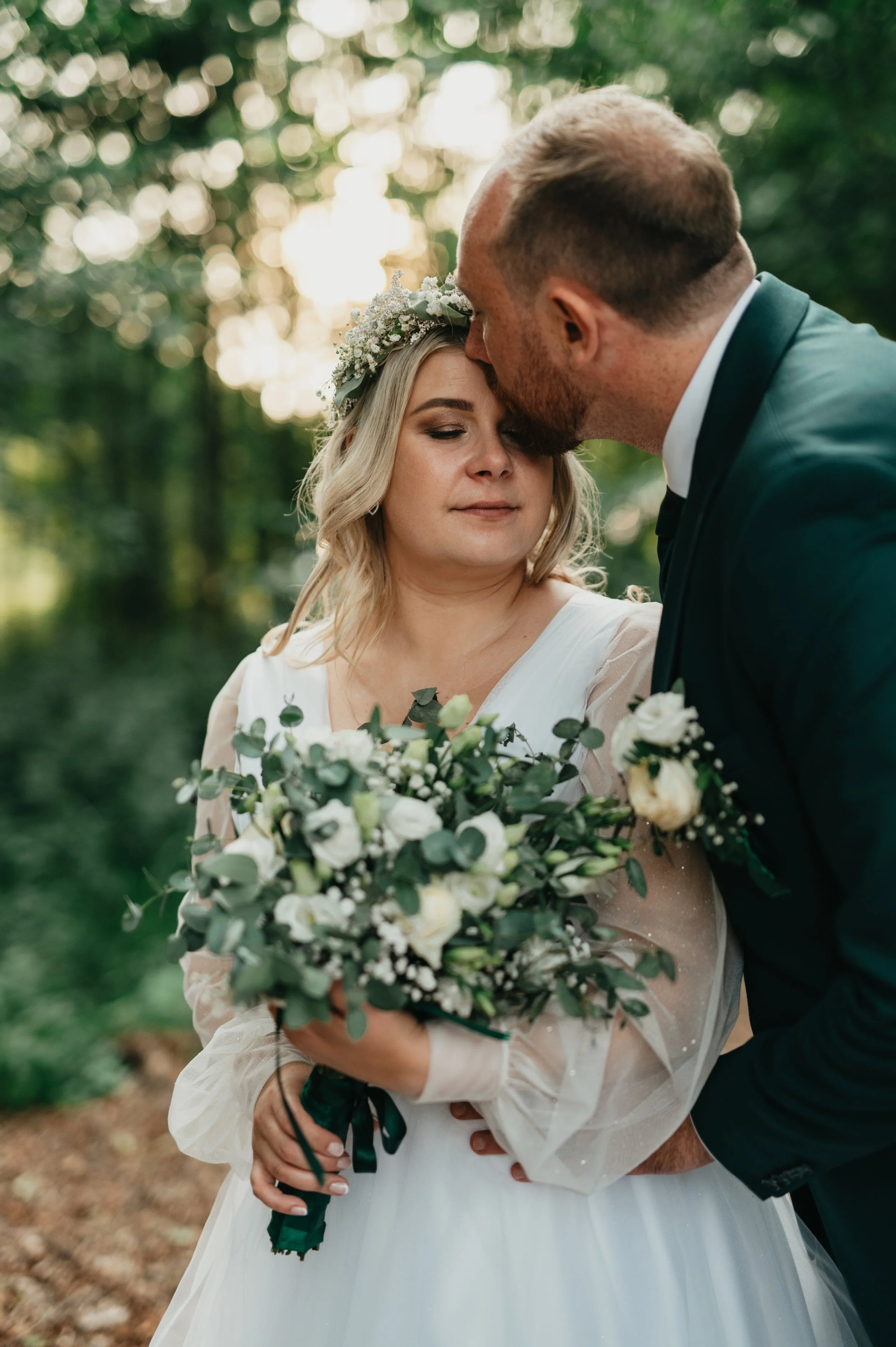 A bride in a white wedding dress with a floral crown and bouquet, and a groom in a dark suit, sharing an intimate moment outdoors surrounded by trees.